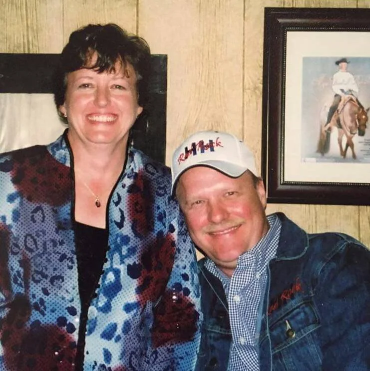 A woman and a man smiling for a photo indoors. The woman has short dark hair and is wearing a colorful patterned jacket. The man is wearing a baseball cap, a checkered shirt, and a denim jacket. There is a framed picture of a person riding a horse on the wall behind them.