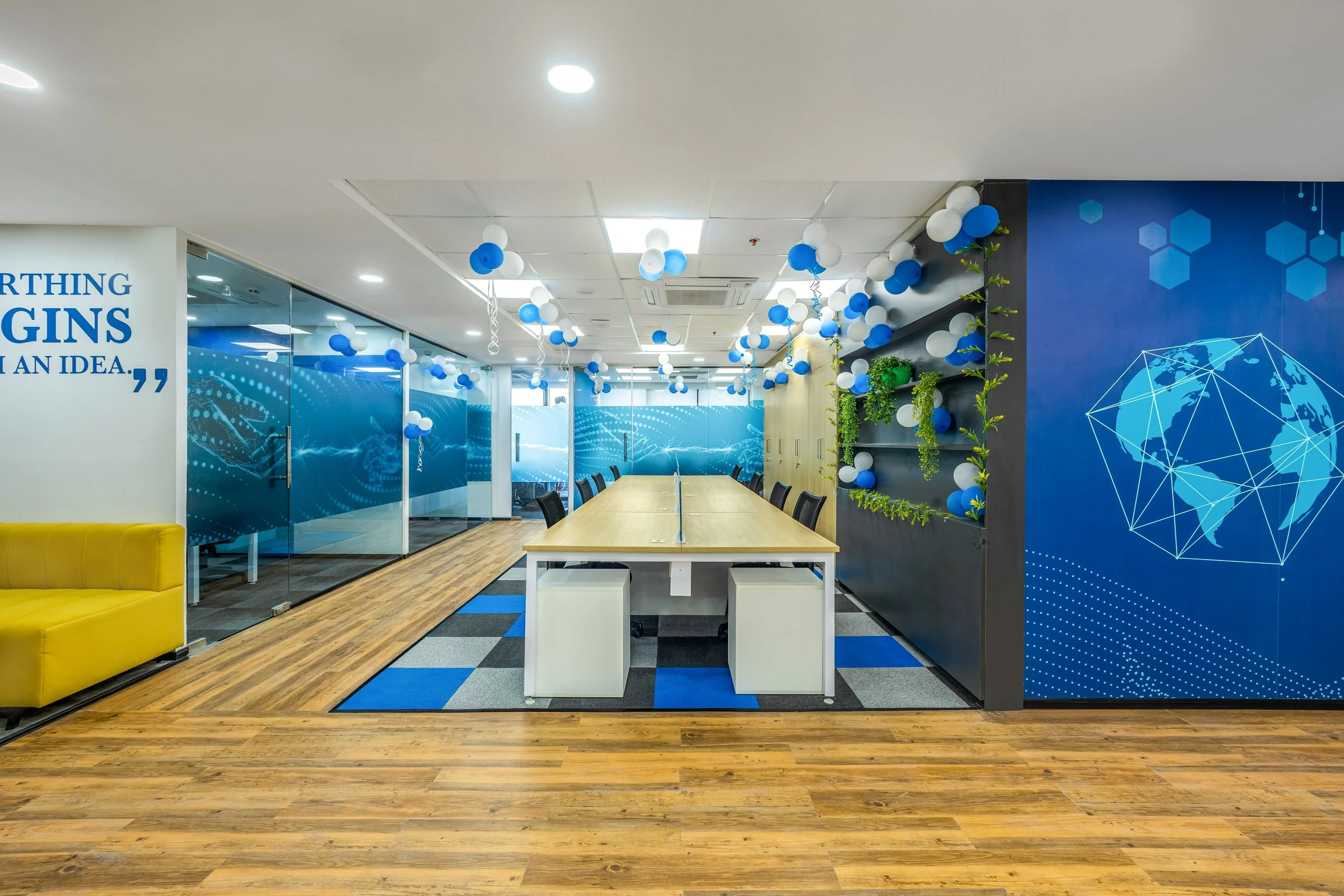 Office conference room decorated with blue and white balloons hanging from the ceiling, with a yellow couch on the left and a long table with chairs, along with a wall featuring a digital globe graphic.