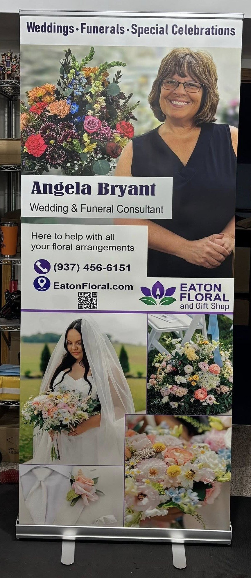 A colorful floral arrangement featuring a variety of flowers such as roses, chrysanthemums, and greenery, next to a smiling woman in glasses with short brown hair. The sign advertises Angela Bryant, wedding and funeral floral and event planning services at Eaton Floral and Gift Shop, with contact information and a website.