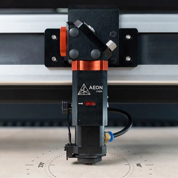 Close-up of a laser engraving or marking machine head with the label 'AEON LASER', positioned above a work surface with measurement markings.