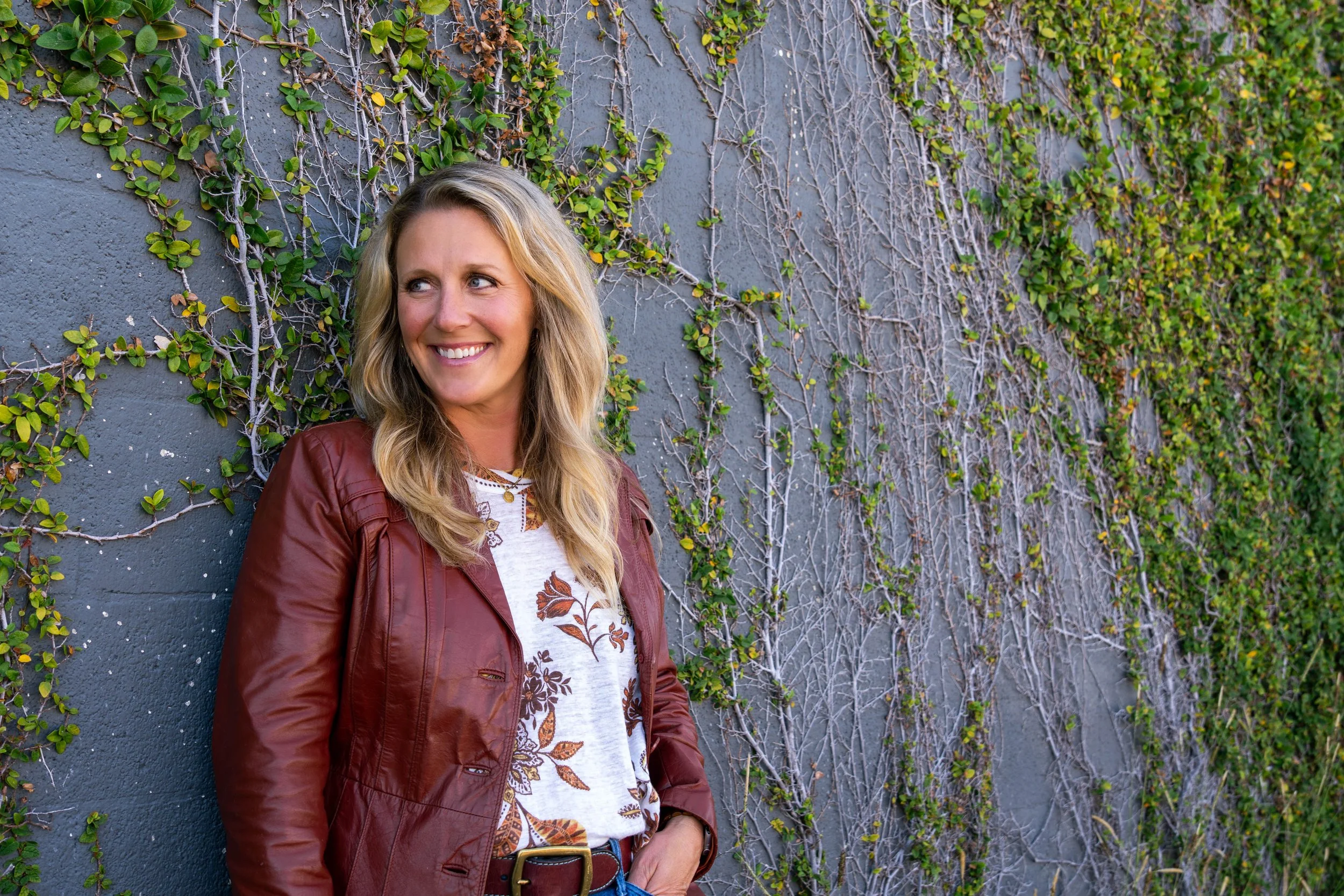 A woman with long blonde hair, wearing a brown leather jacket and patterned shirt, smiling and leaning against a gray wall covered with green and brown ivy.