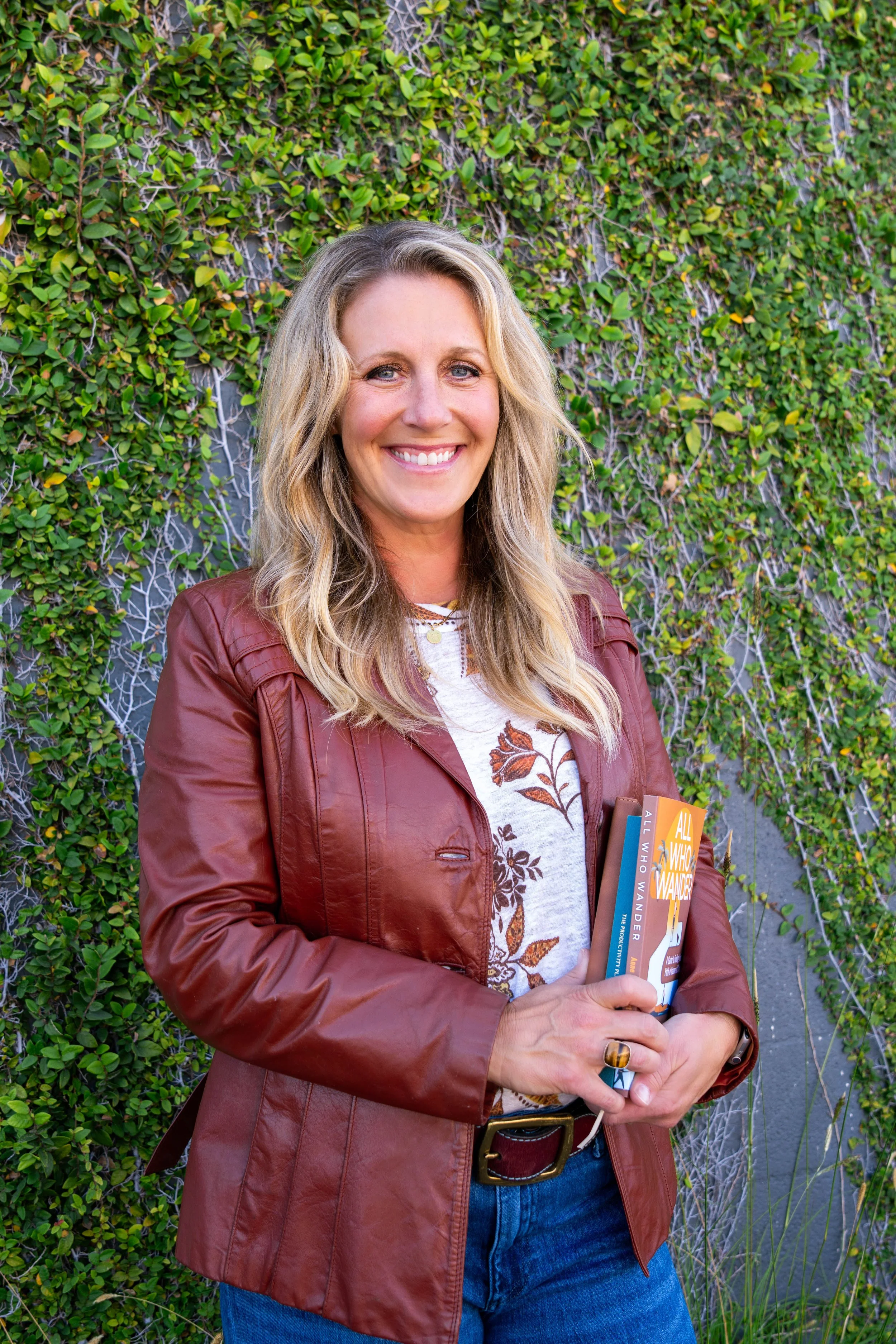 A smiling woman with blonde hair wearing a maroon leather jacket, blue jeans, and a white graphic t-shirt holding books standing in front of a green leafy wall.