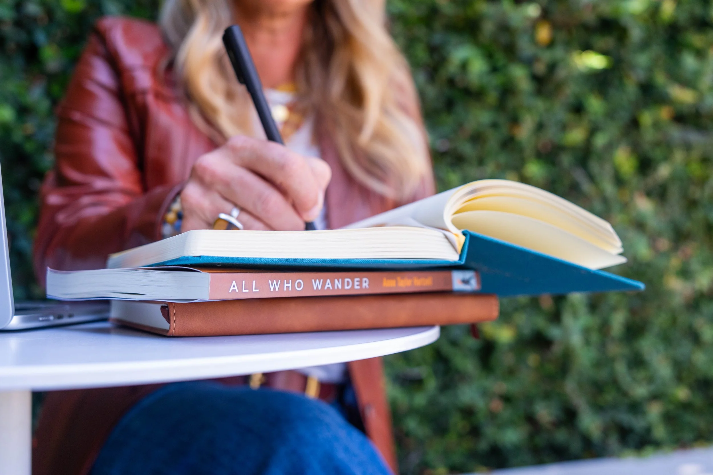 A person sitting at a white table outdoors, writing in a notebook with a black pen. There are three books on the table, one titled 'ALL WHO WANDER'. The person is wearing a brown jacket and has long blond hair. The background shows green foliage.