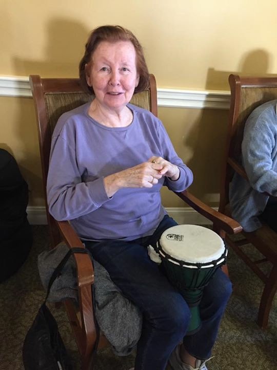 An elderly woman with short red hair and a light purple long-sleeve shirt sitting on a wooden chair, holding a small drum with a white top, in a room with beige walls and a patterned carpet.