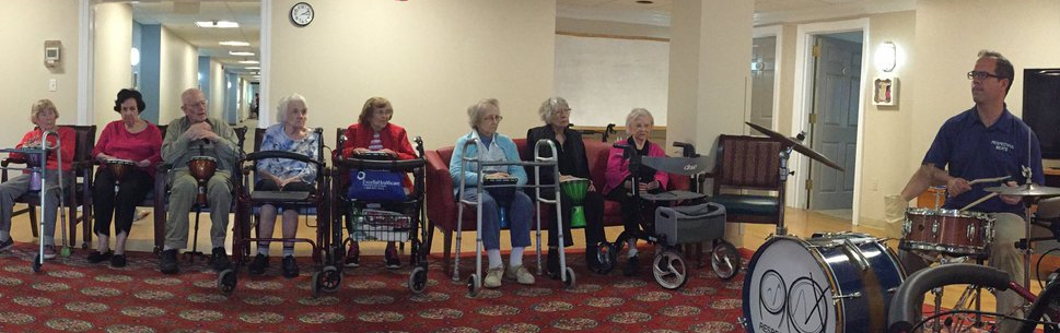 Group of elderly people seated in a row in a common area, with a music therapist playing drums in front of them.