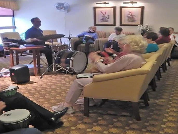 A group of elderly people sitting in a living room, listening to a musician playing drums and cymbals.