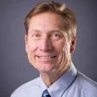 A middle-aged man with light brown hair, smiling, wearing a light blue shirt against a gray background.