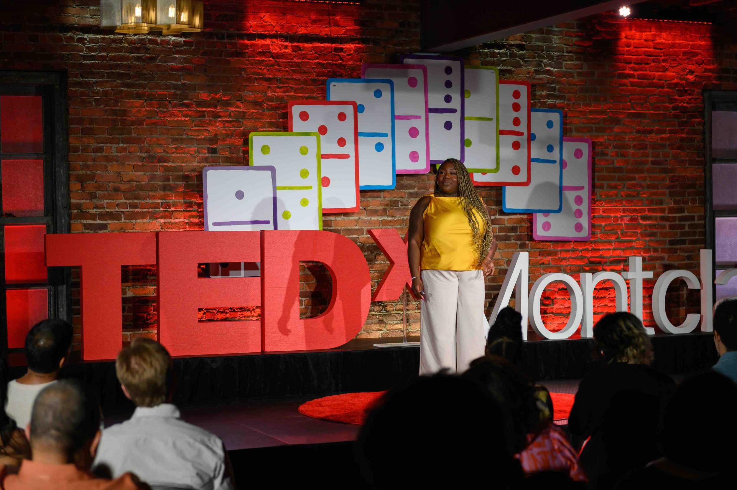 Speaker in a yellow top and white pants on stage in front of the TEDx Montclair sign