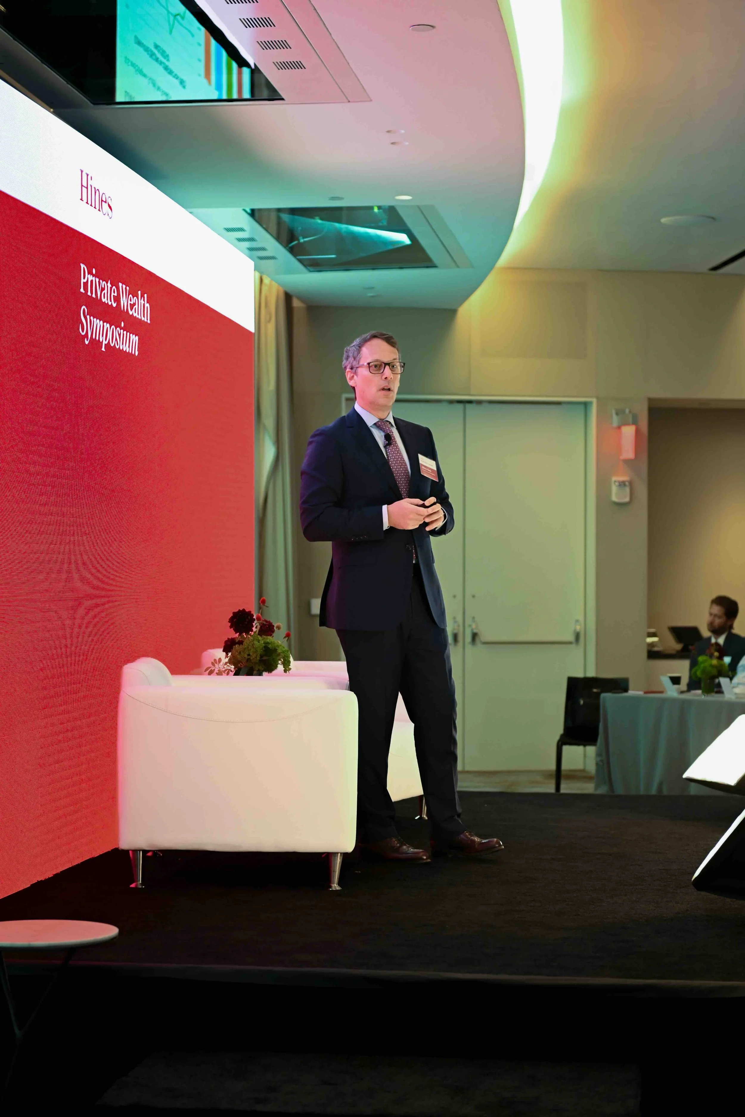 Speaker standing near white lounge chairs on stage at the Hines symposium