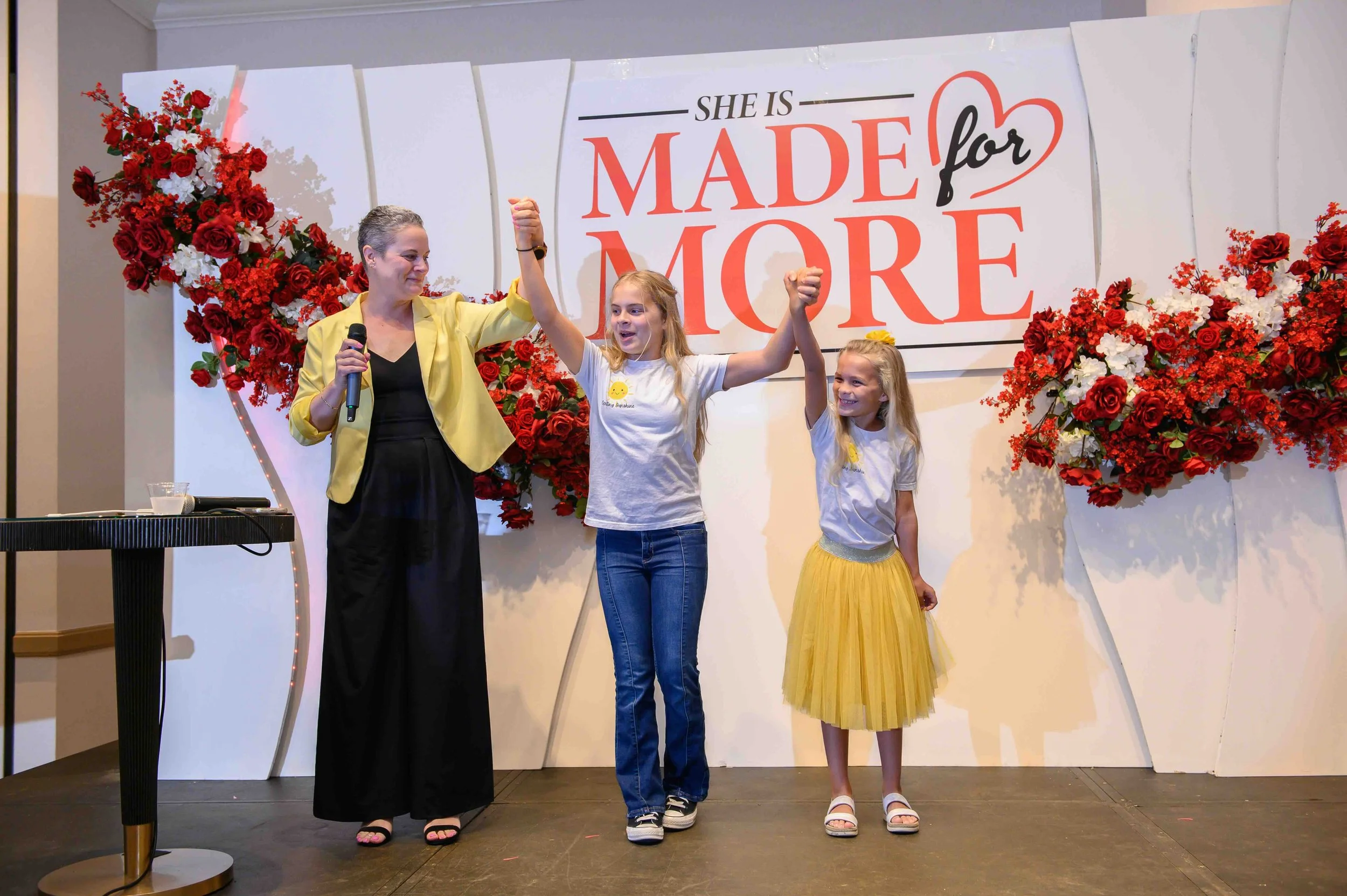 Three women cheering on stage in front of the floral backdrop