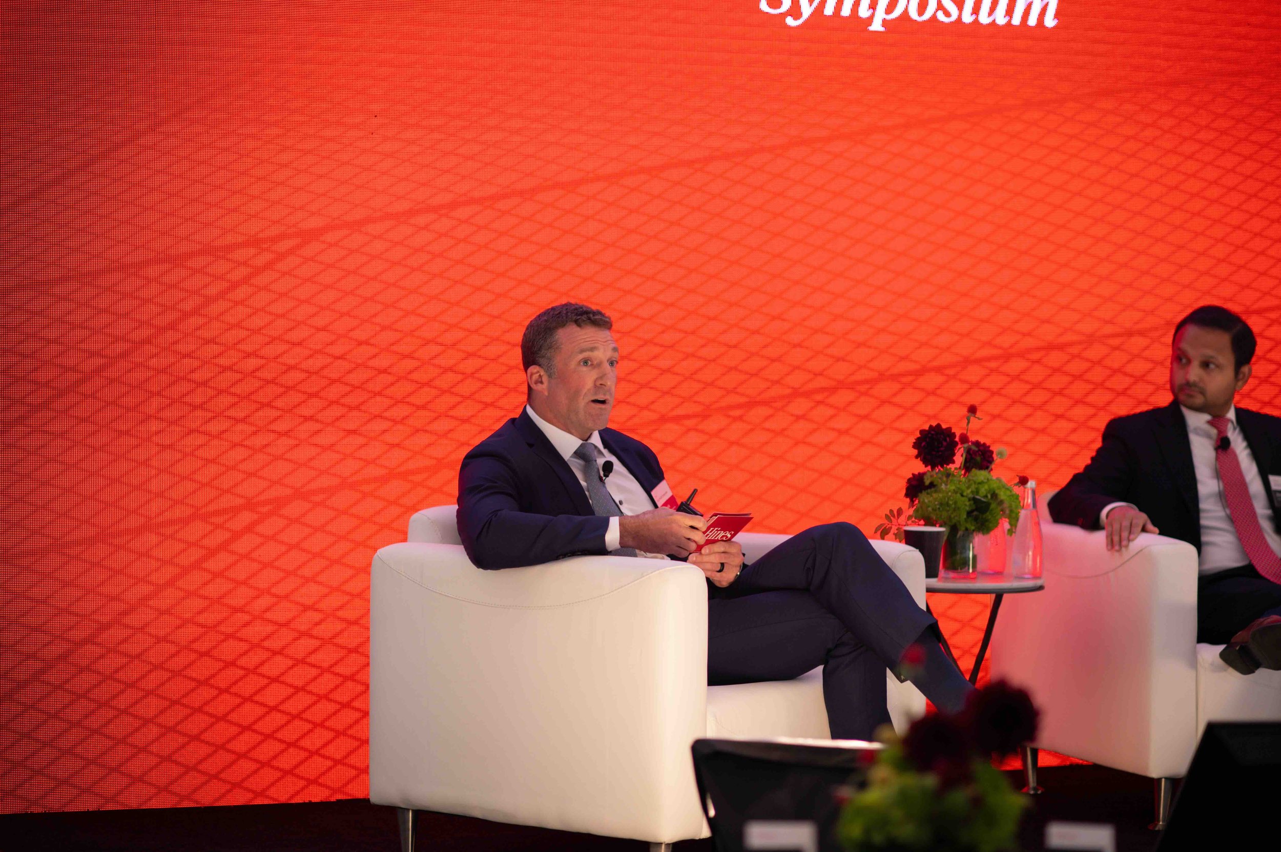 Speaker seated in a white chair on stage in front of a red backdrop at the Hines symposium