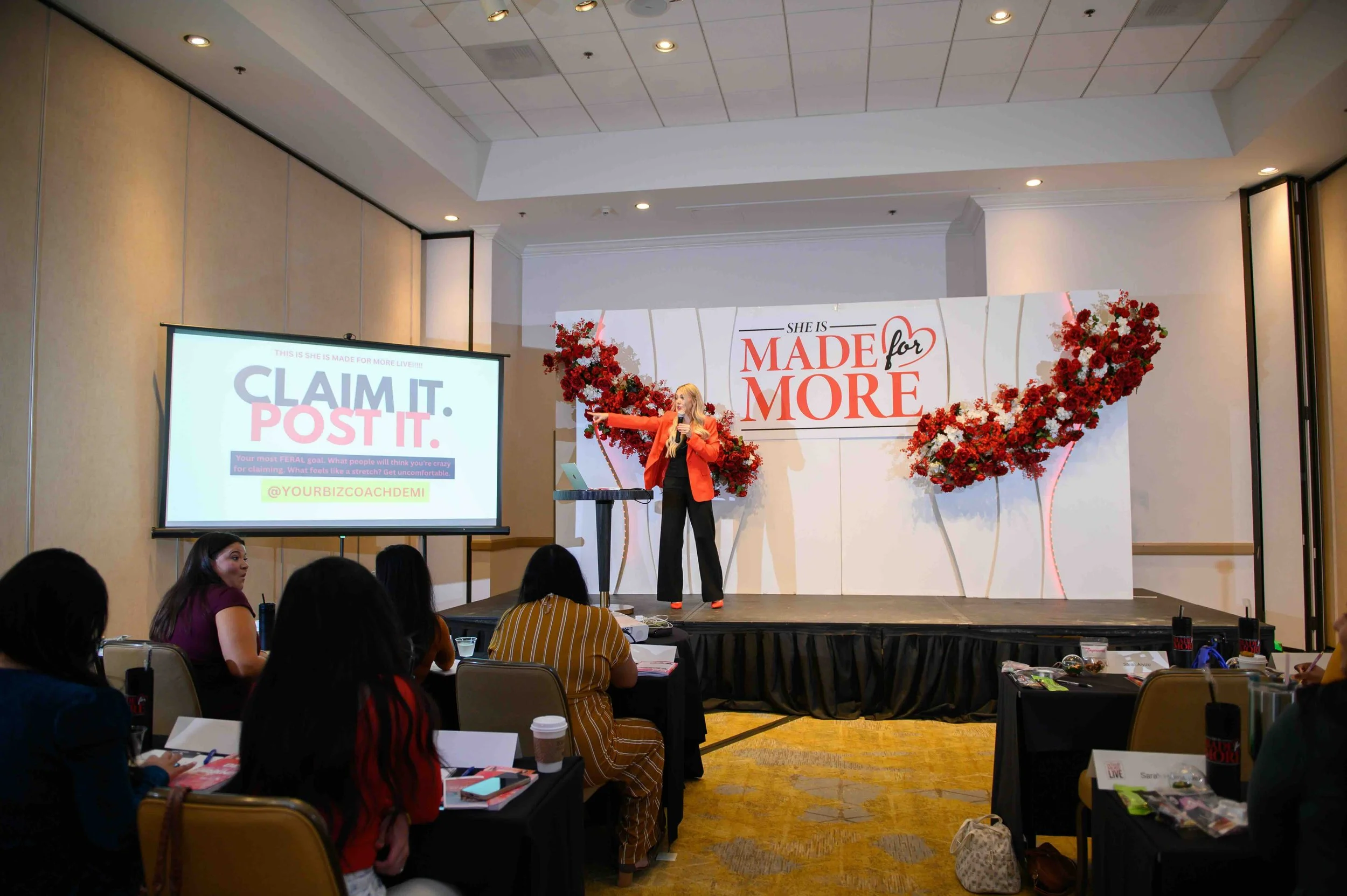 Wide view of attendees seated at tables with a presentation screen and floral backdrop