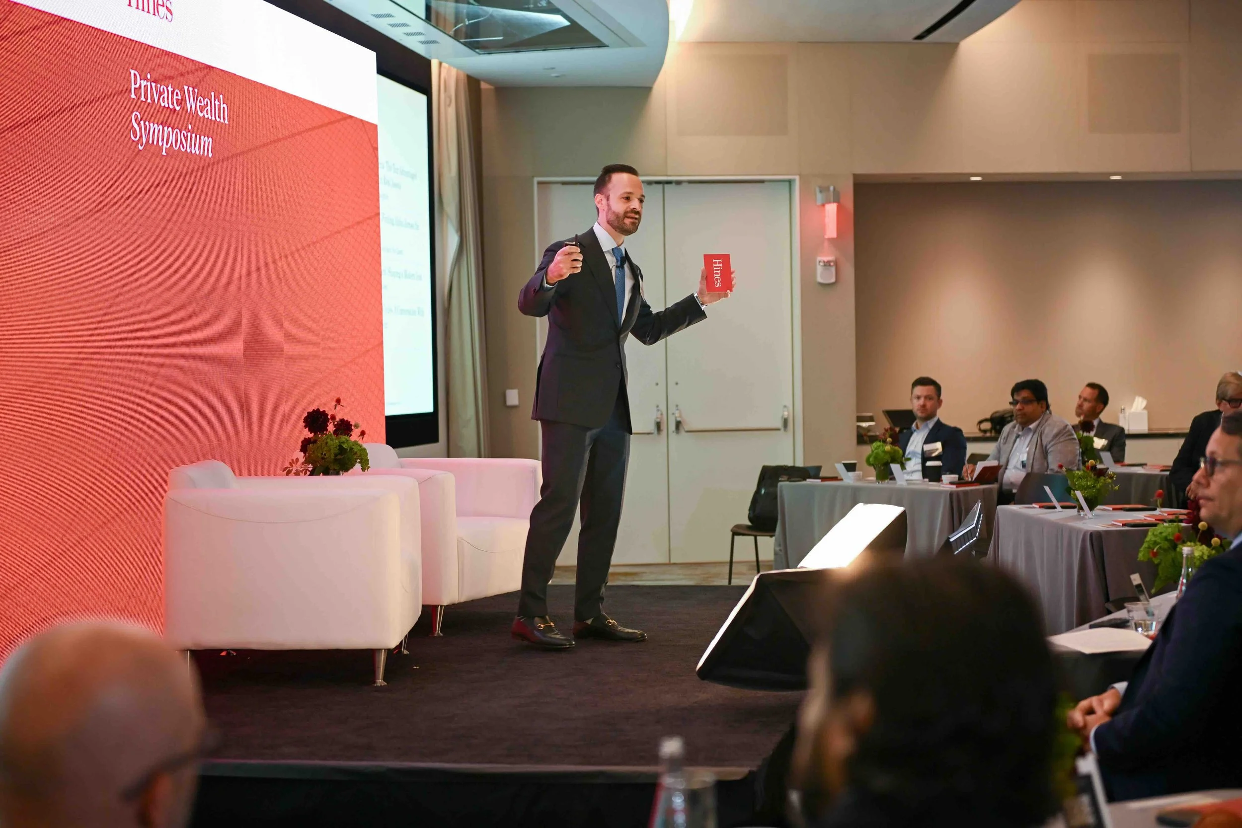 Speaker in a suit presenting beside a red wall at the Hines symposium