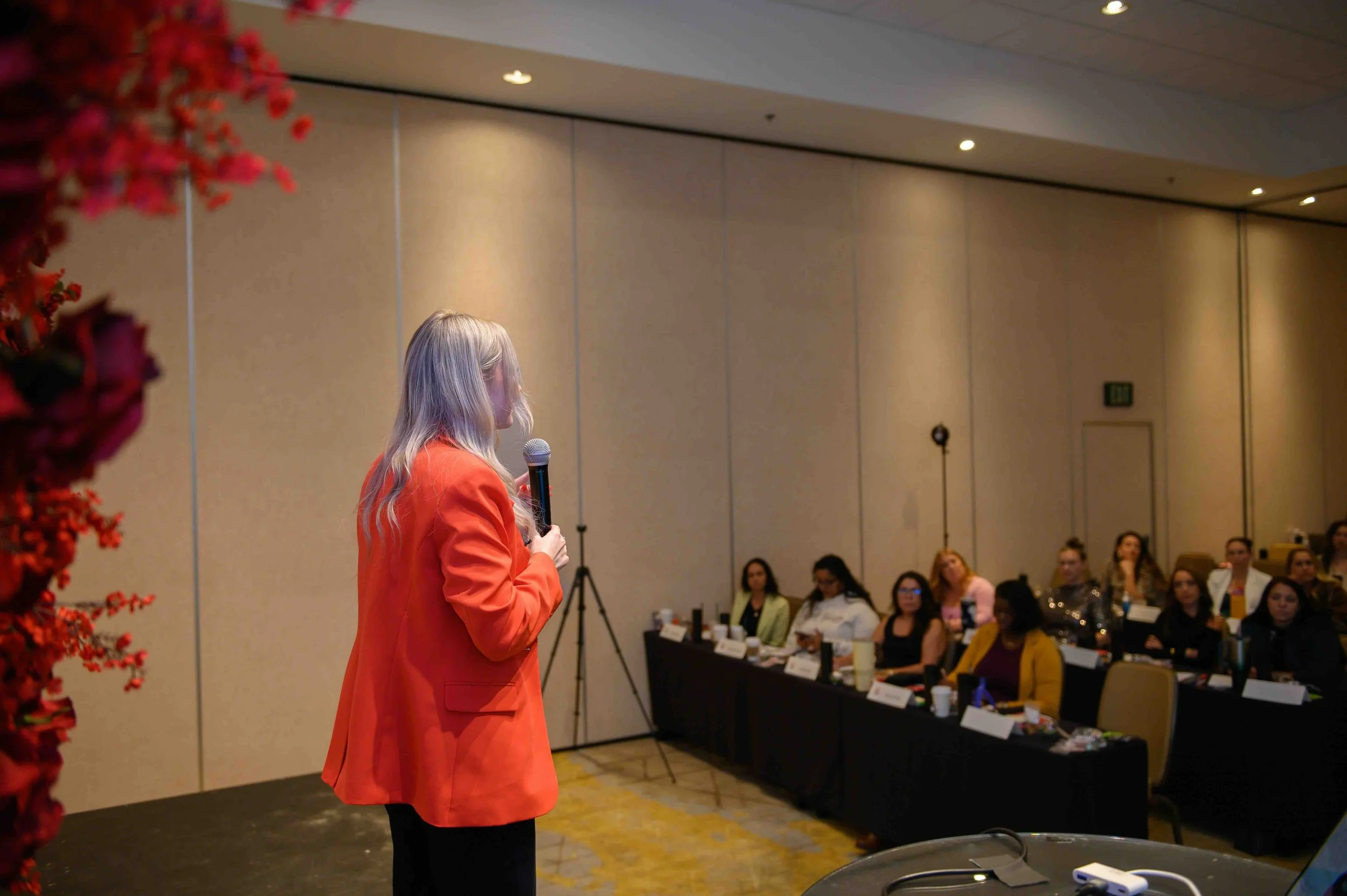 Speaker in an orange blazer holding a microphone and presenting to the audience