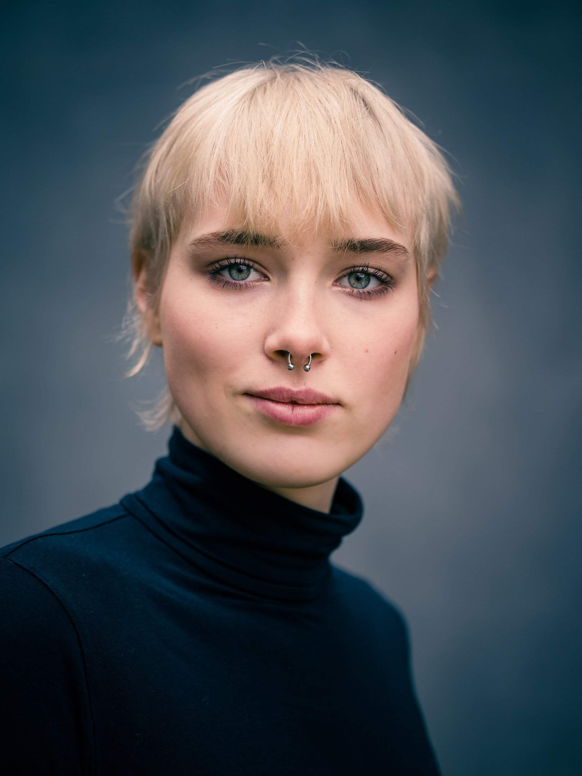 Porträt einer Person mit kurzem, blondem Haar und Nasenpiercing, die ein schwarzes Oberteil trägt.