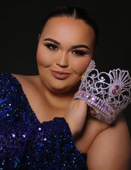 A woman with dark hair and bold makeup, wearing a dark blue sequined dress, holding a decorative crown with gemstones in front of her face against a dark background.