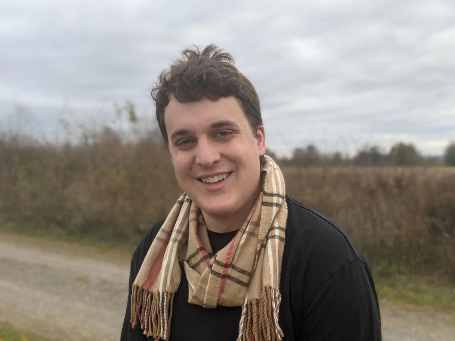 Young man with curly brown hair smiling outdoors, wearing a black jacket and a beige plaid scarf, against a cloudy sky and rural landscape background.