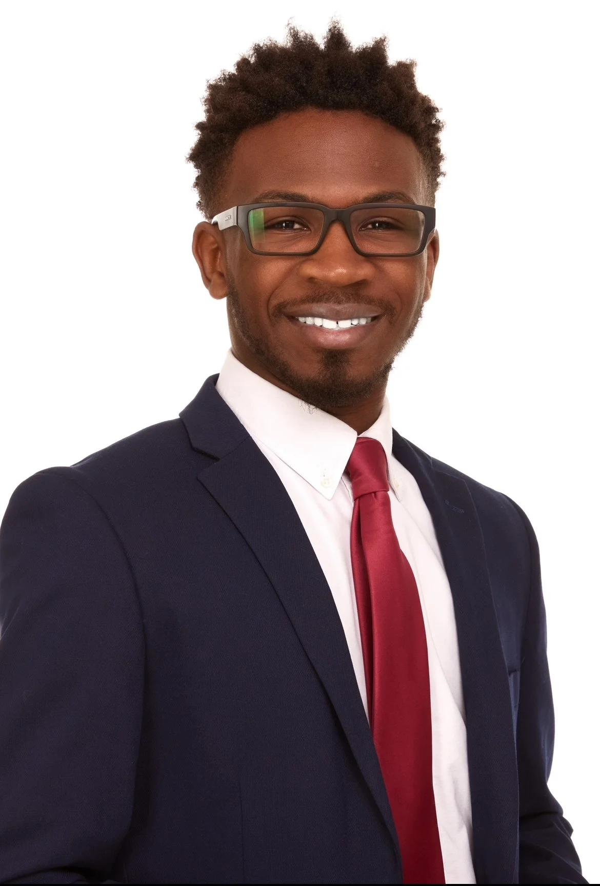 A young Black man in a navy suit, white shirt, and red tie, wearing glasses, smiling against a plain white background.