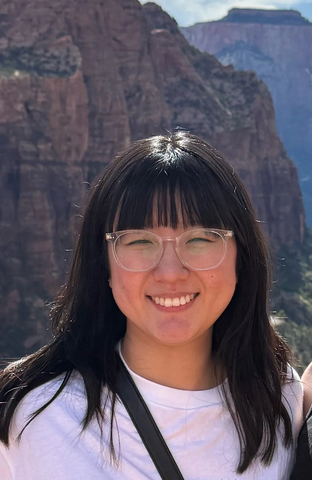 A woman smiling with glasses, dark hair, standing in front of a large canyon landscape with rock formations and cliffs.