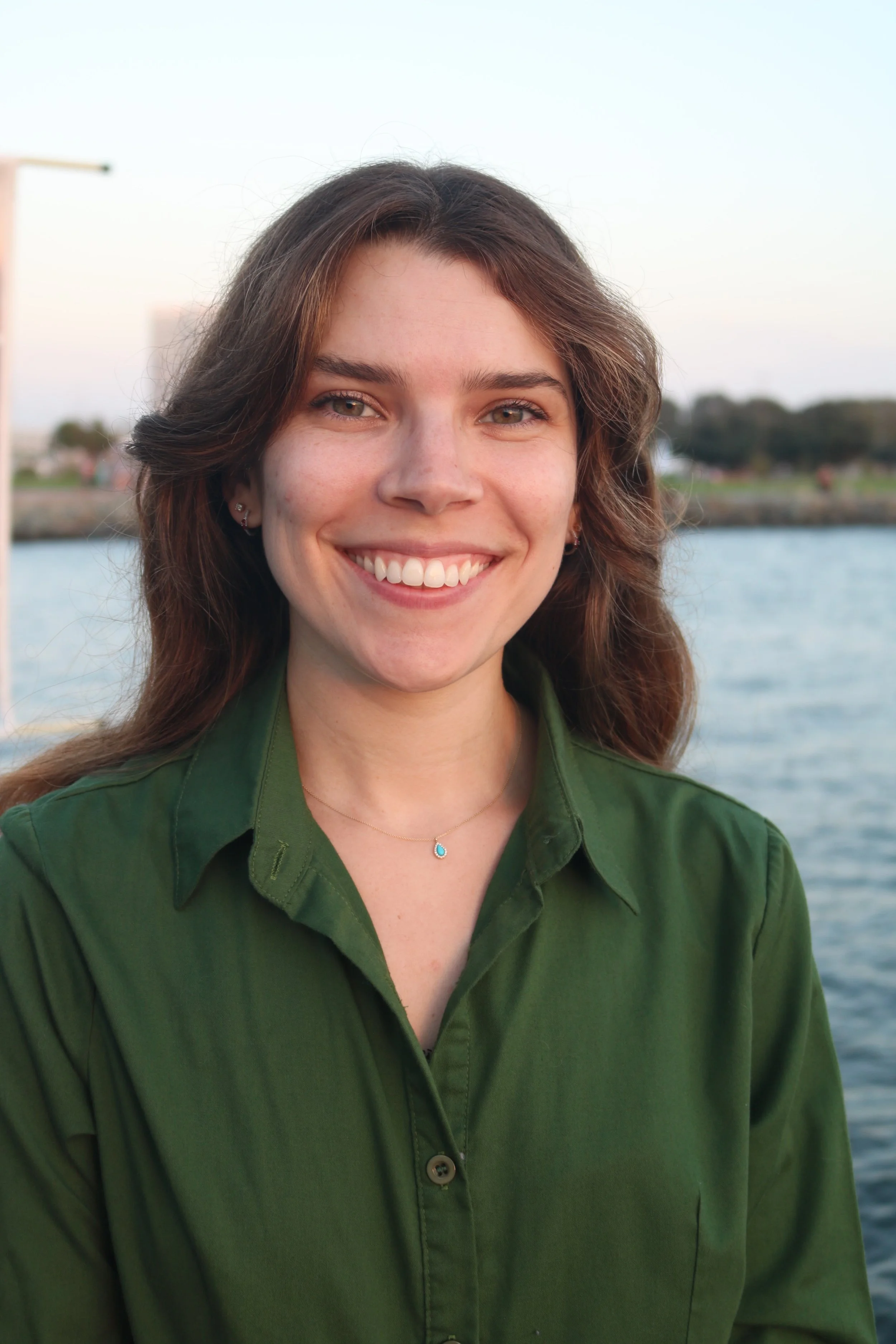 A smiling young woman with brown hair wearing a green shirt standing outdoors near water at sunset.