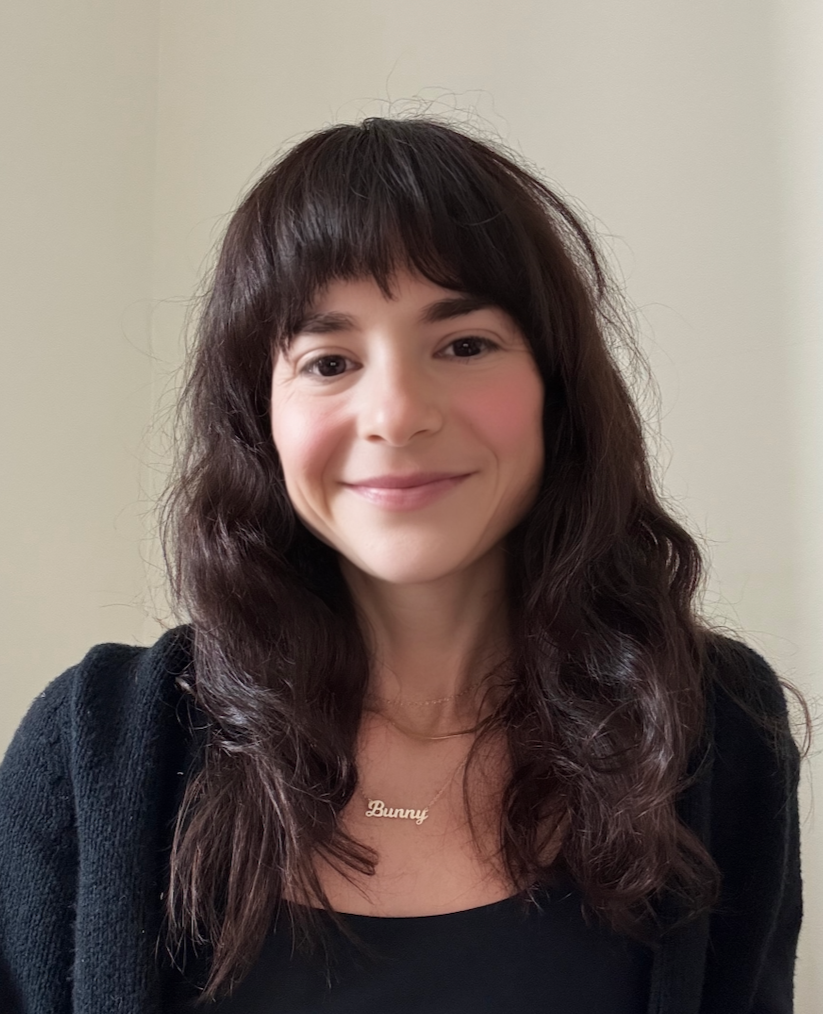 A woman with long, wavy dark hair and bangs smiling at the camera, wearing a black top and a gold necklace that says 'Bunny'.