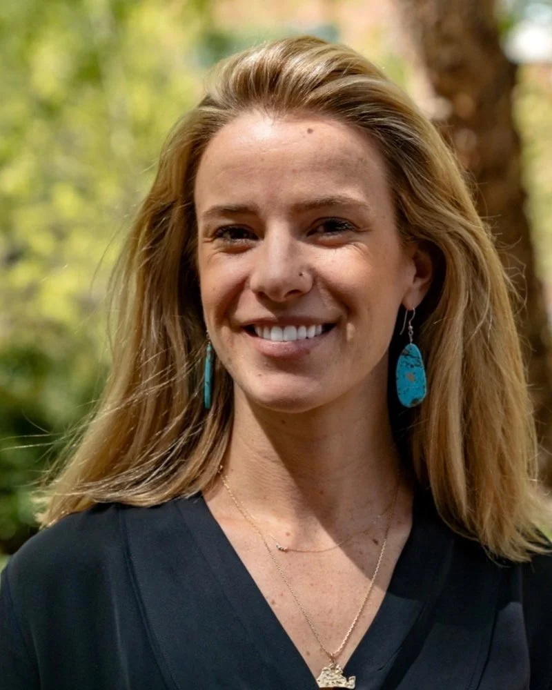 A woman with long, light brown hair smiling outdoors, wearing turquoise earrings and necklaces, with blurred trees in the background.