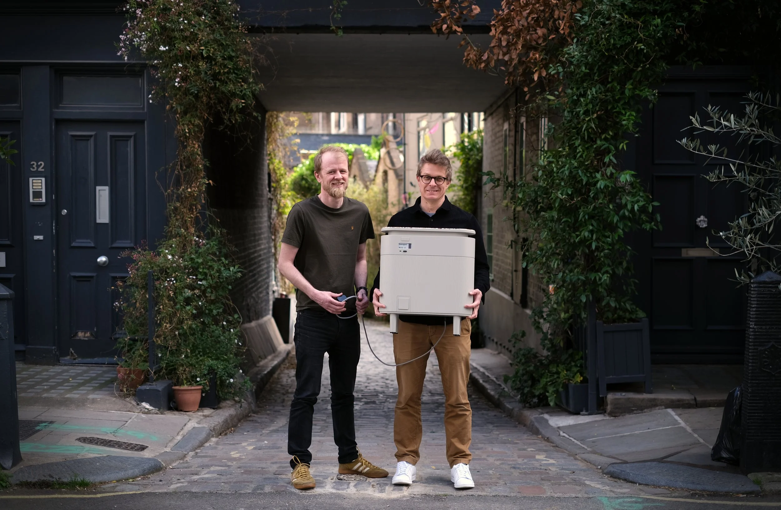 Ashley and Rob, founders of Windfall Energy, pose outdoors on a London street holding a plug-in home battery.