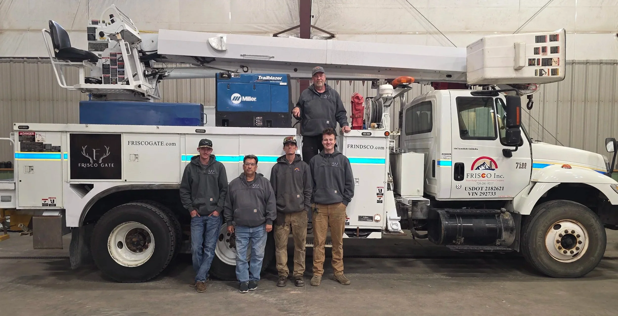 A group of five men standing in front of a white utility truck with a tall extended boom lift inside a warehouse or garage. The men are wearing gray hoodies with a logo, some with glasses and hats, and are smiling or looking serious.