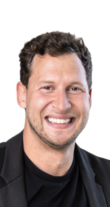 A man with short, curly brown hair smiling and showing teeth, wearing a black shirt and dark jacket.