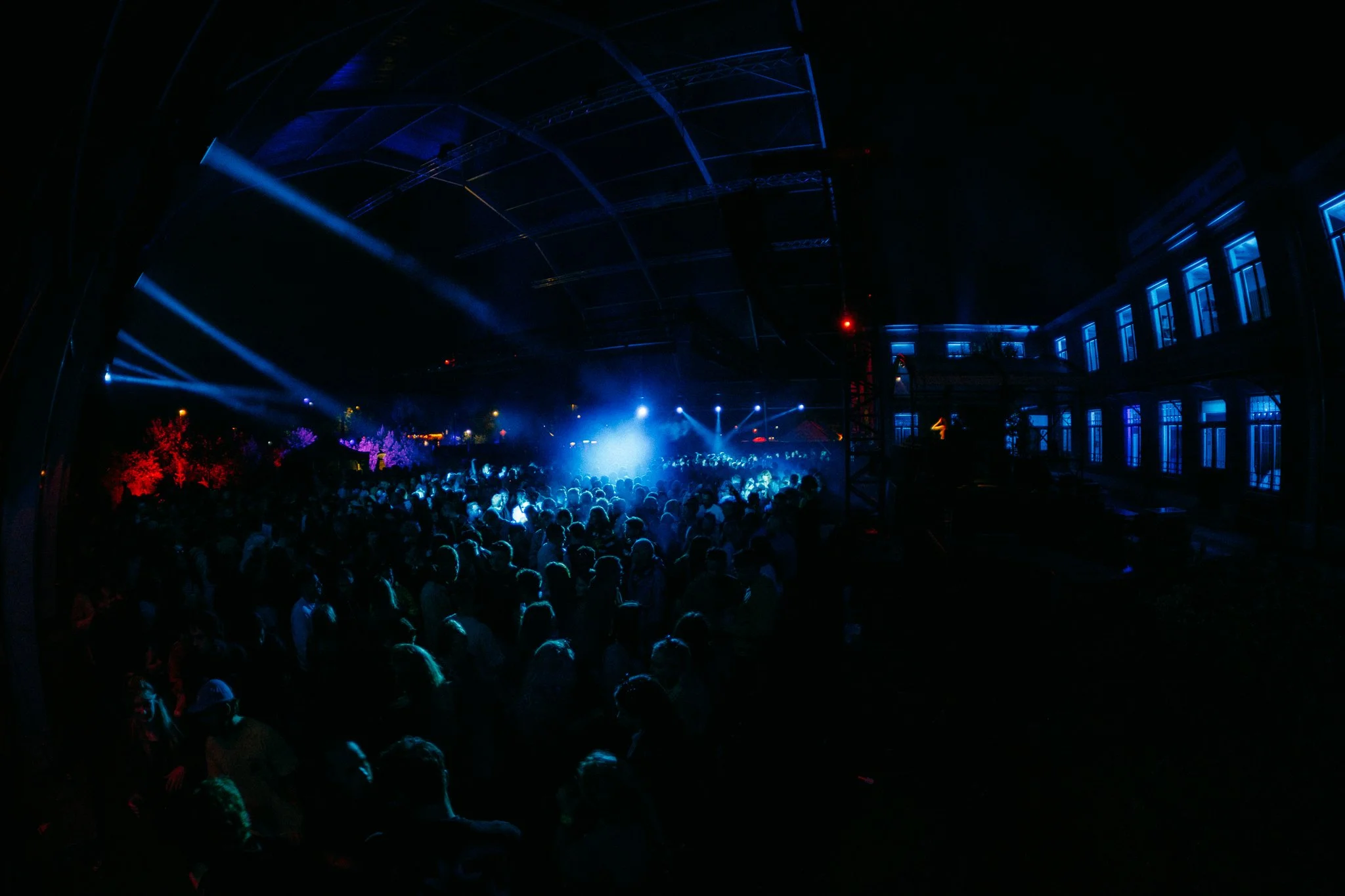 Een druk dancefeest met blauwe en paarse lichten onder een grote open-structuur, mensen dansen en genieten.