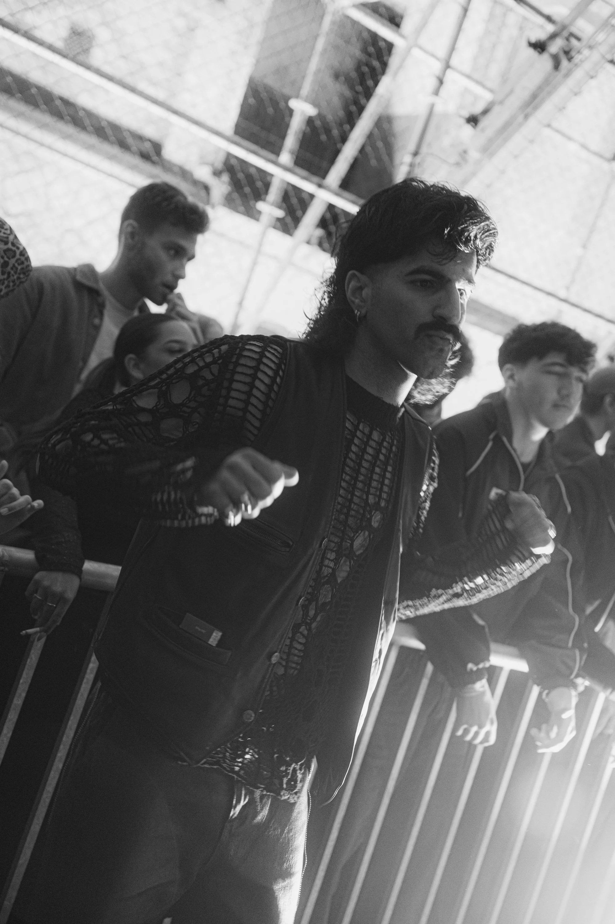 Man in zwarte jas met opengewerkte mouwen en legerbroek, omringd door mensen, onder een baldakijn in zwart-ww.