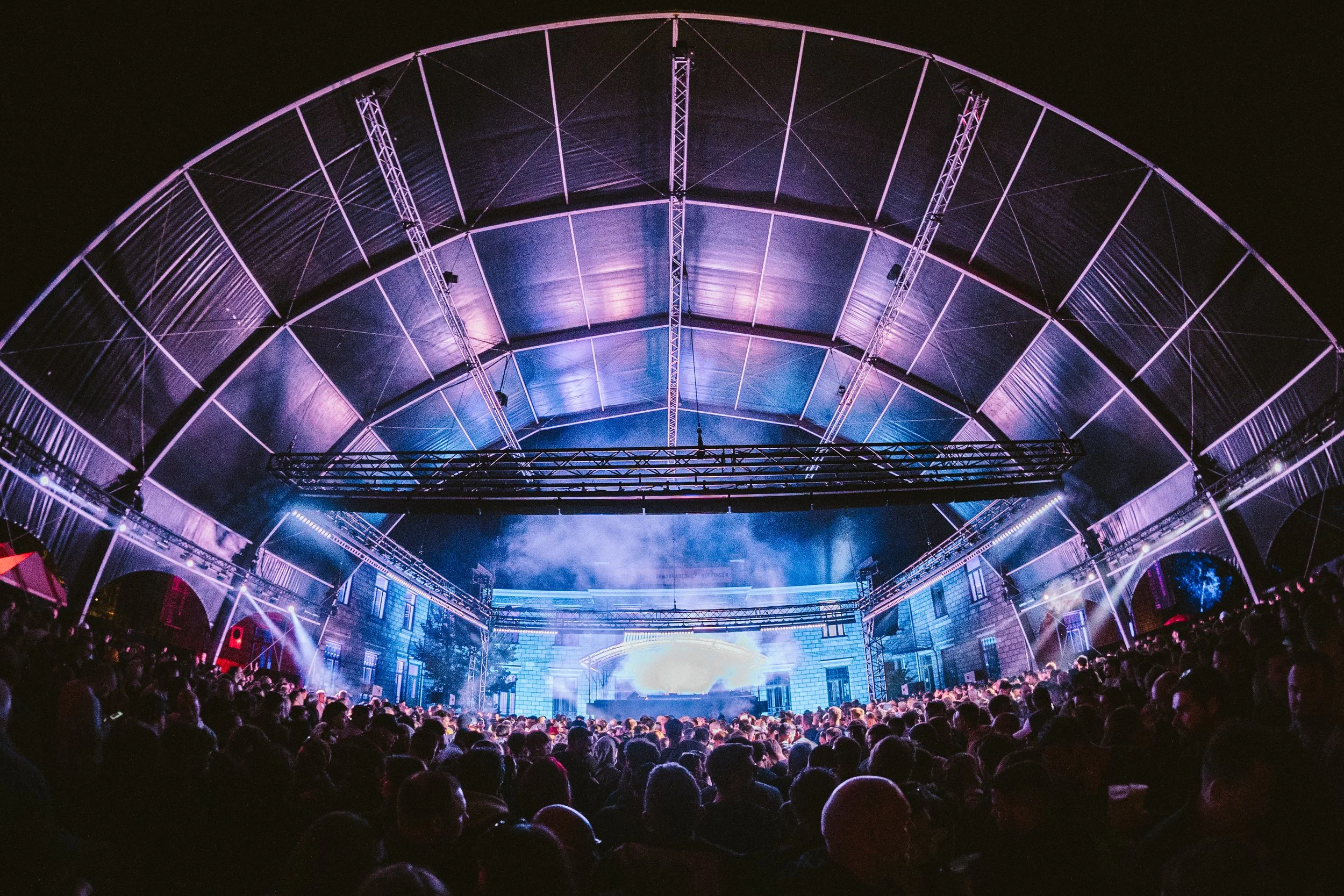 Een grote menigte mensen kijkt naar een verlichte podium onder een grote, transparante tentstructuur tijdens een nachtelijke muziek- of cultuurwereldvoorstelling.