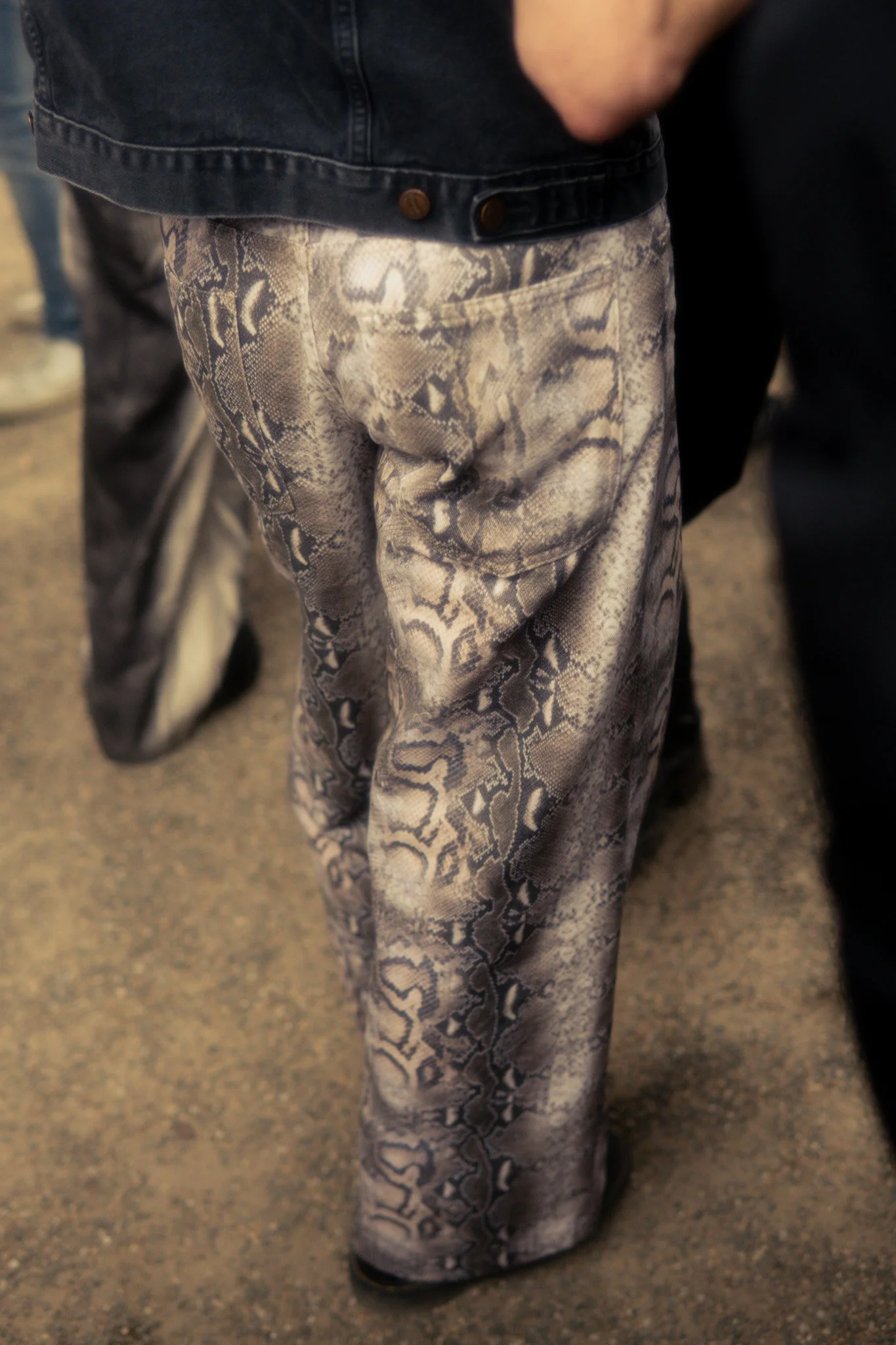 Close-up van een persoon met beige en grijze slangenprintbroek en een donkerblauwe jas.