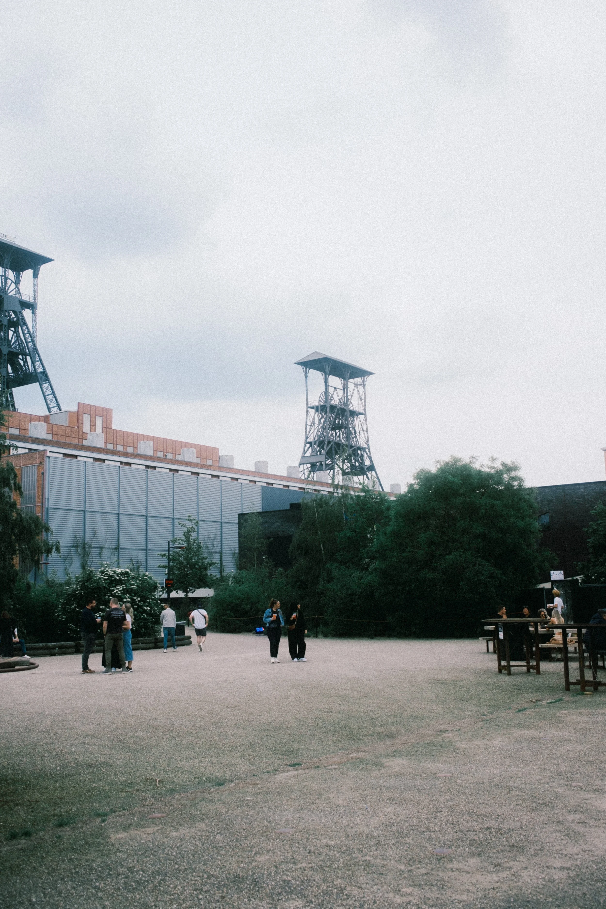 Openbare plek met mensen die wandelen en zitten, met industriële structuren en groene bomen op de achtergrond, onder een bewolkte lucht.