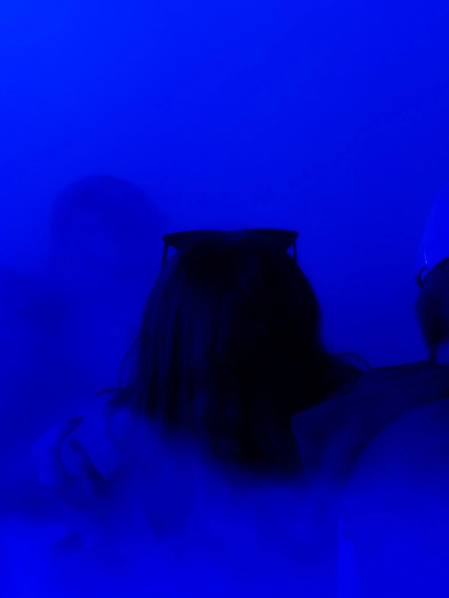 Twee mensen zitten in een donkere kamer met blauw licht, de persoon in het midden heeft een zonnebril op de neus.