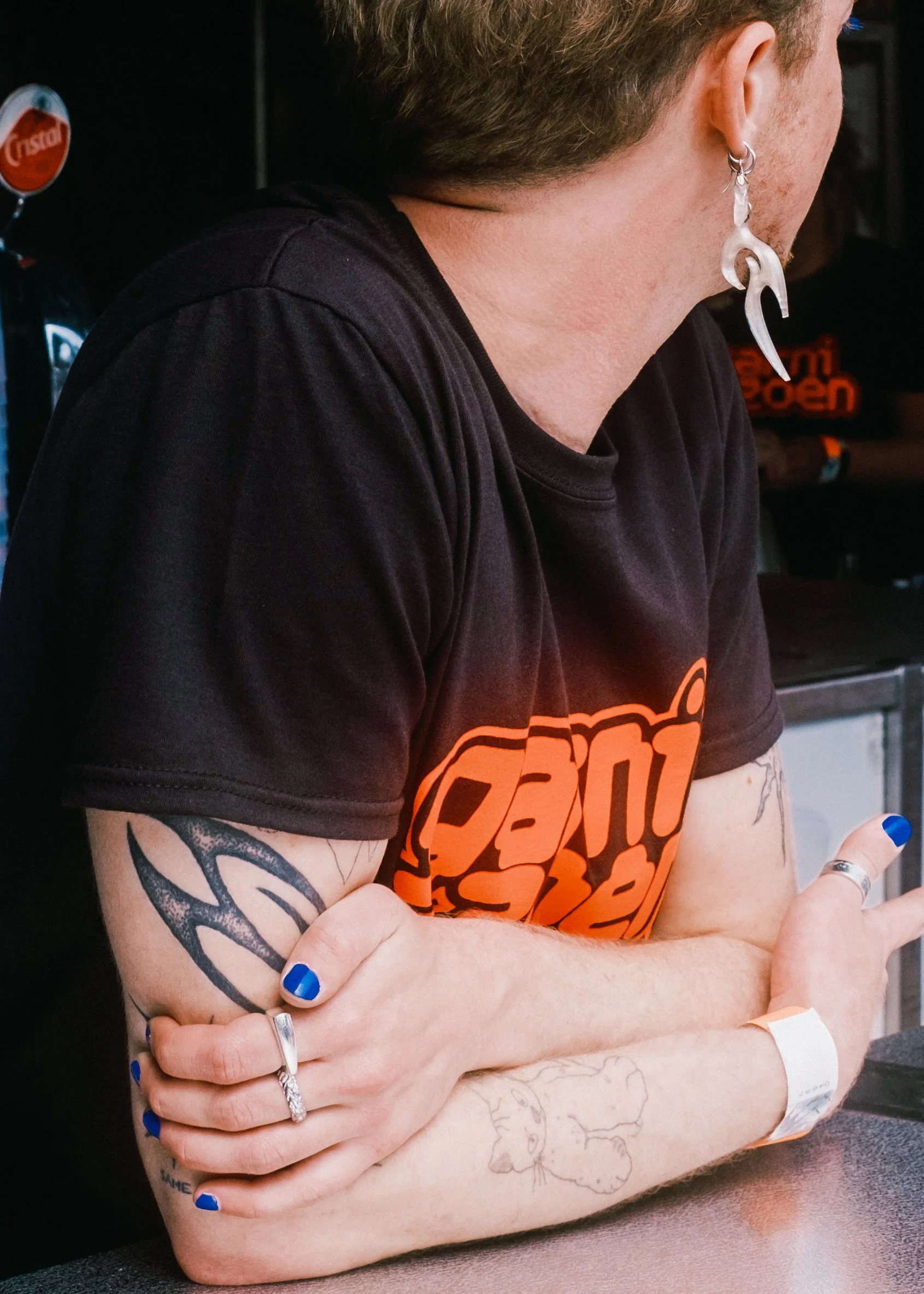Close-up van een persoon die met gekruiste armen op een tafel zit, met opvallende blauwe nagels, tatoeages en sieraden, dragend een zwarte T-shirt met een oranje logo, en met oorbellen en andere sieraden.