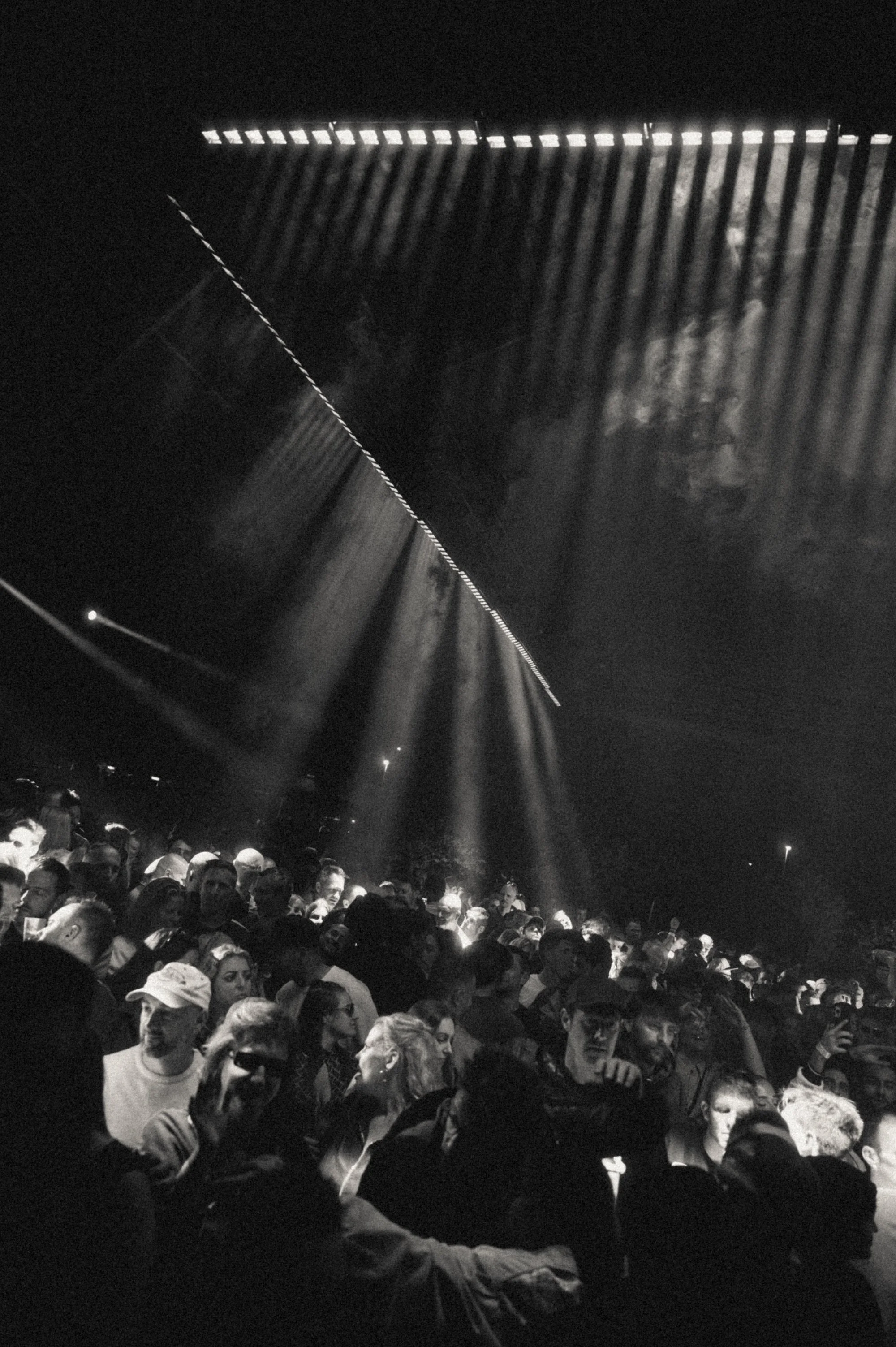een druk festivalpubliek onder lichtstralen in een donkere omgeving, zwart-wit foto