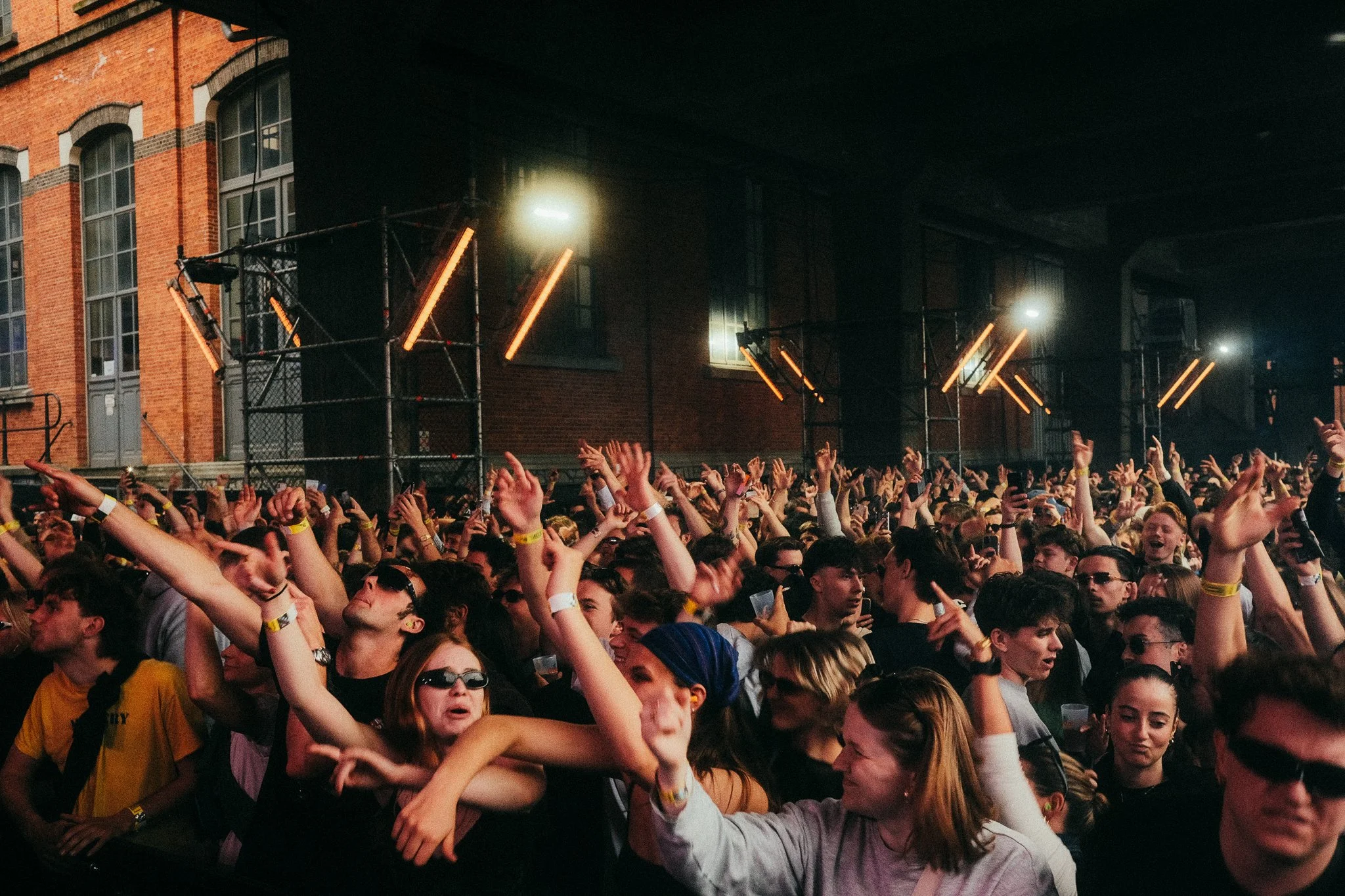 Een grote groep mensen die naar een concert of feestje kijken, met handen omhoog en dansen, binnen een industrieel gebouw met rode bakstenen muren en verlichting.