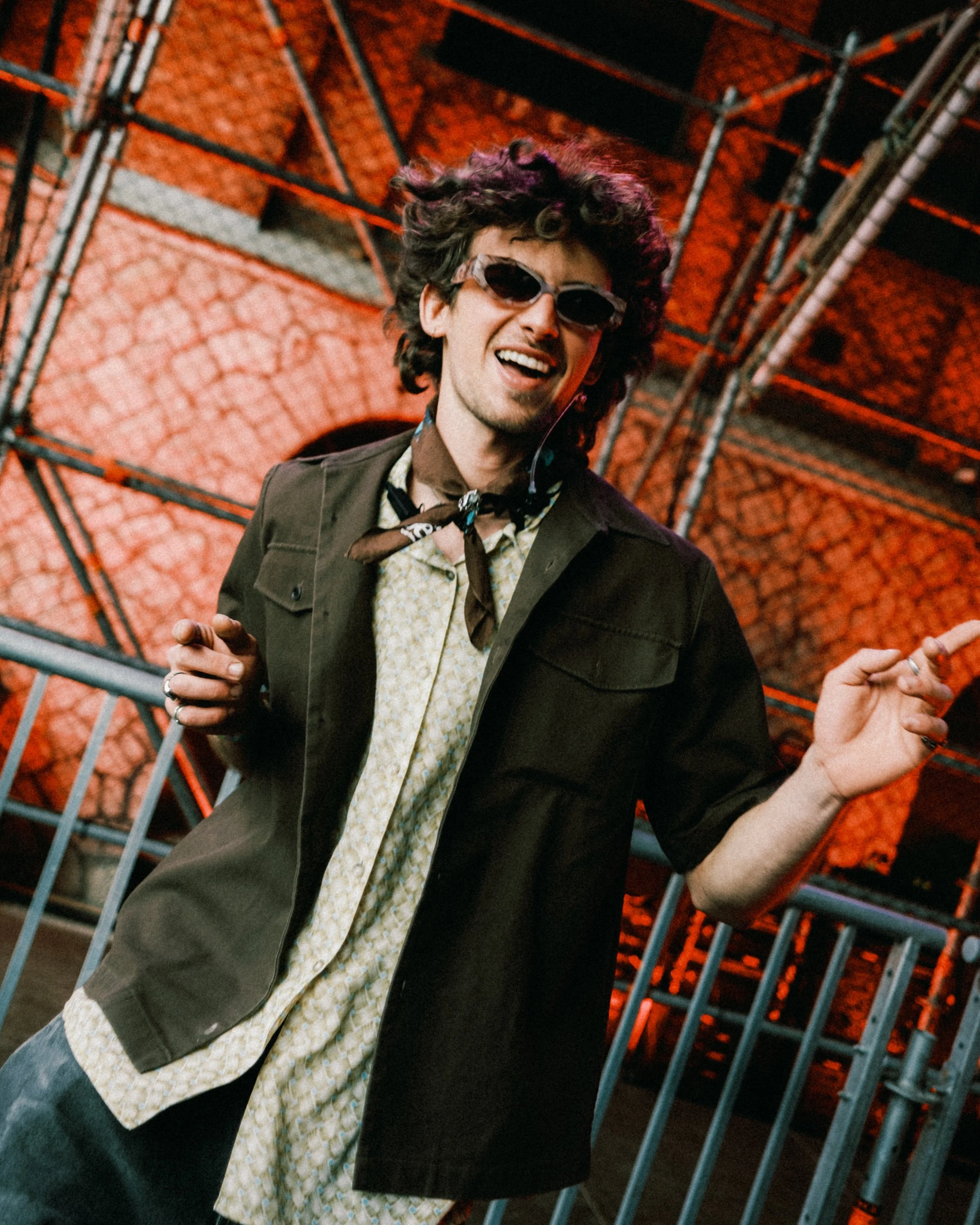 Man met donker gekleurd jack, een lichte blouse en een satijnen sjaal, lachend met zonnebril en dansend of plezier hebbend voor een metalen hek met een rode bakstenen achtergrond.