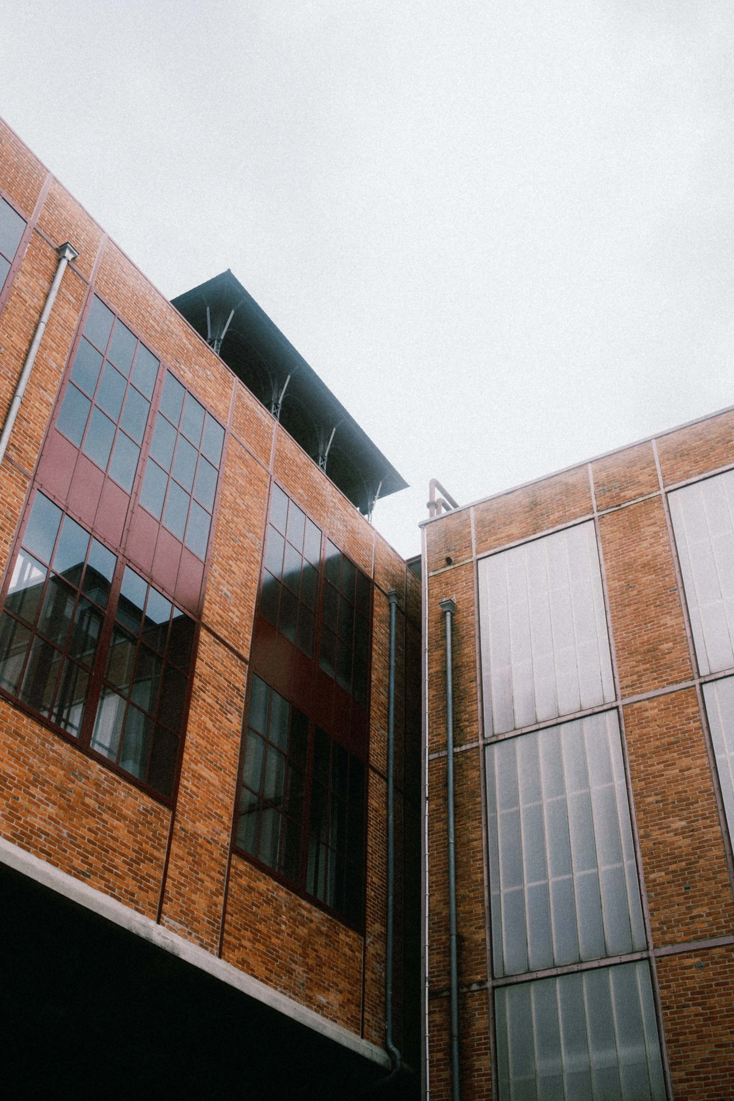 Close-up van een modern gebouw met ruitramen en een overhangdakterras, gemaakt van bakstenen en glas, onder een grijze hemel.