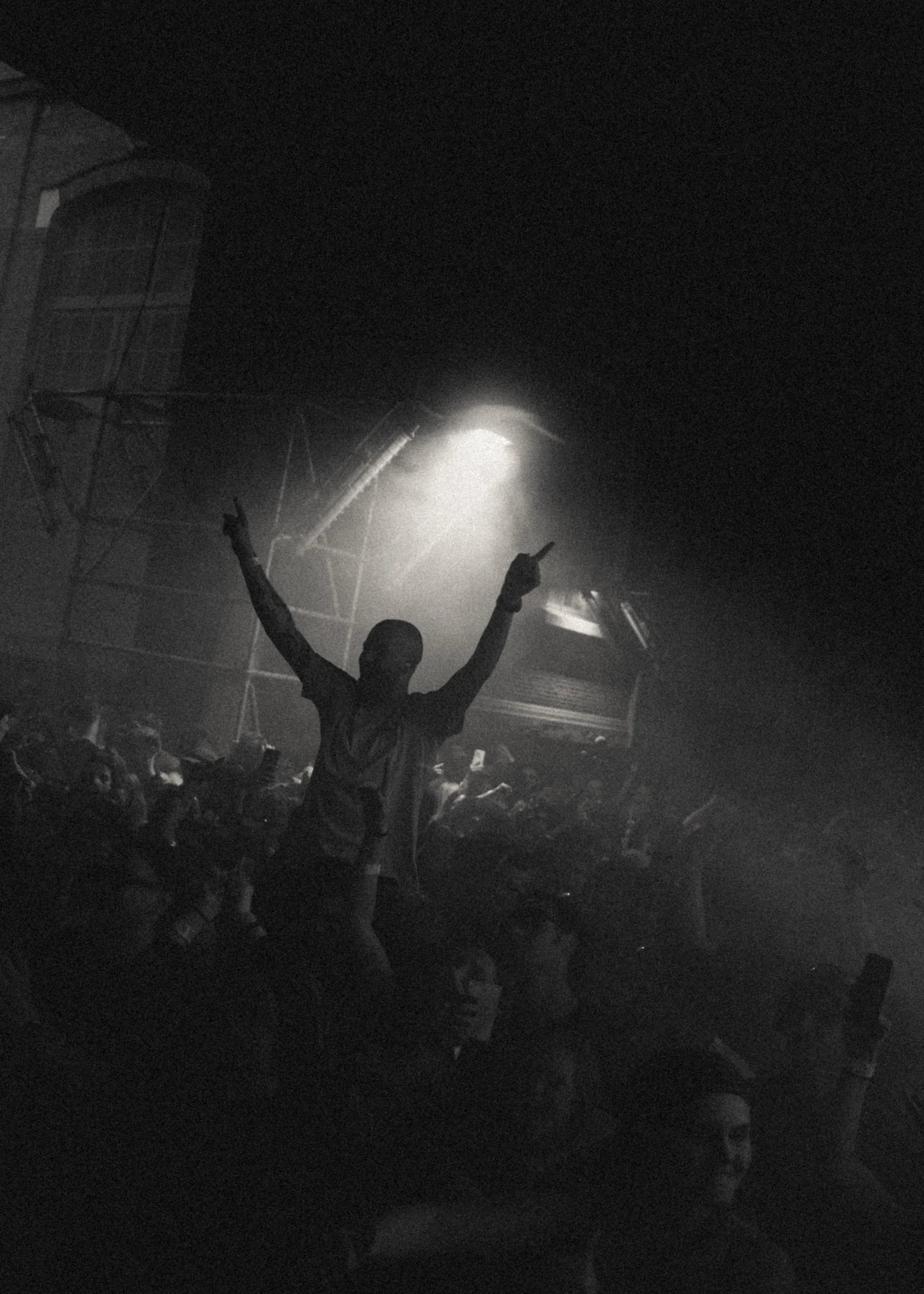 Een persoon met uitgestrekte armen en vingers omhoog in een donkere, drukke menigte tijdens een concert of feest, met een enkele lichtstraal boven hen.