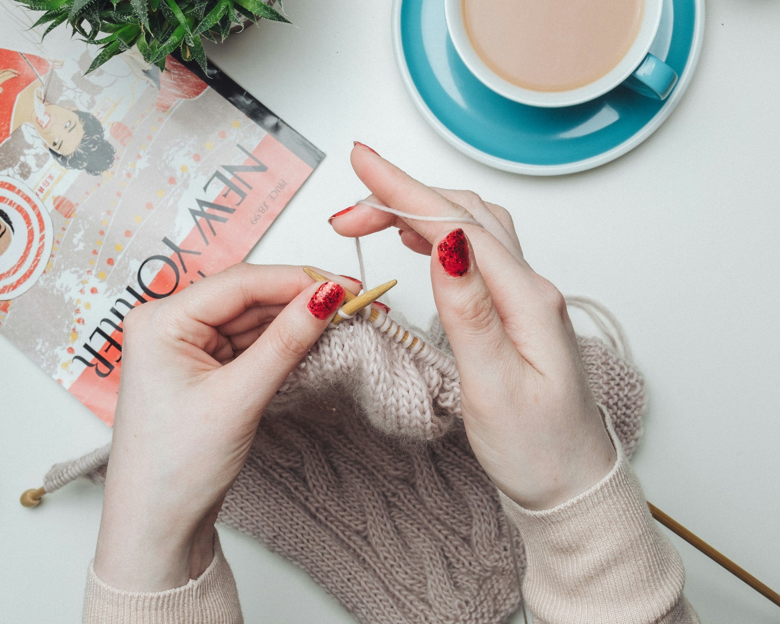 Person knitting with beige yarn, red glitter nail polish, on a white table with a magazine, a blue and white cup of coffee, and a green plant.