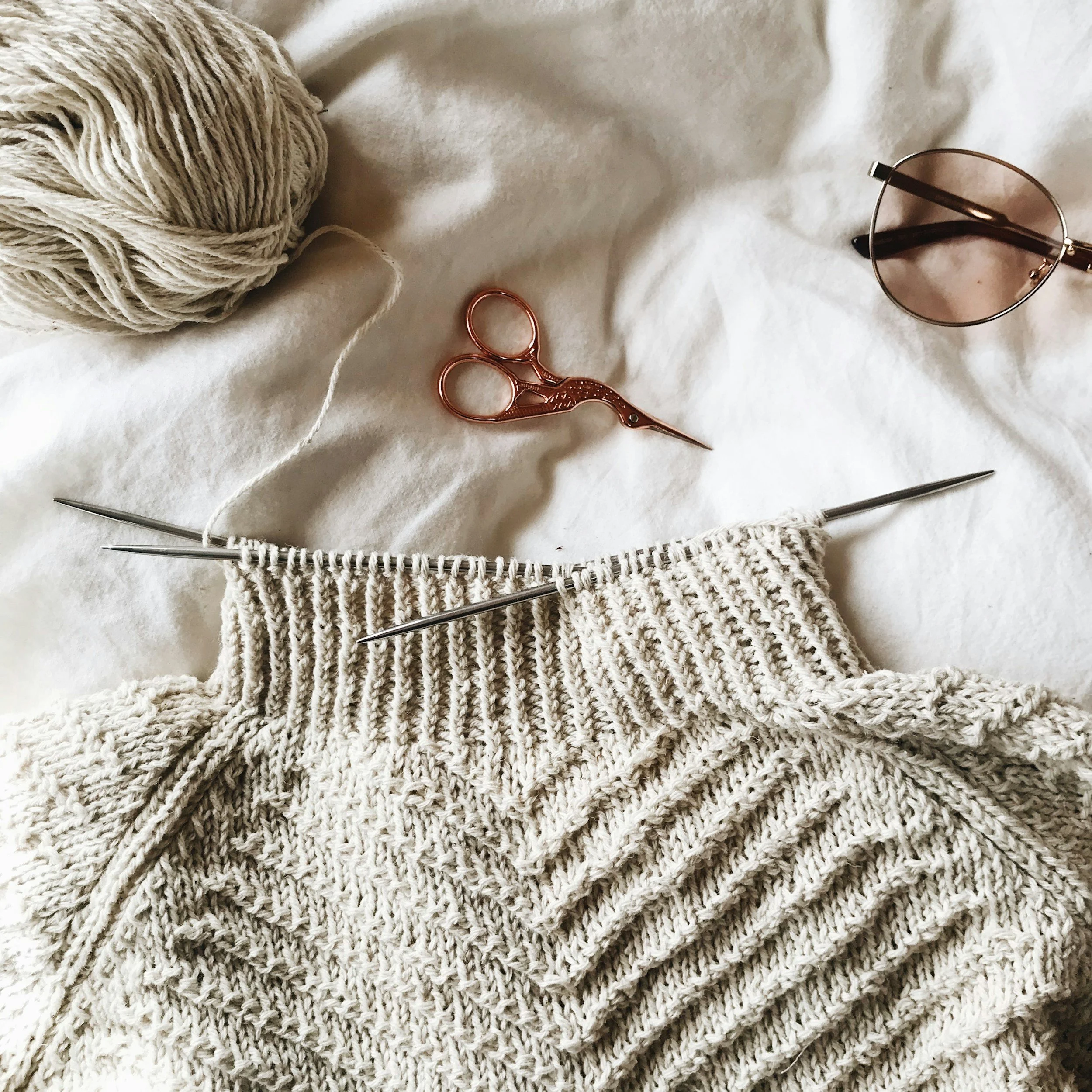 A knitting project in progress with cream-colored yarn, knitting needles, pink-tinted sunglasses, small scissors, and a ball of yarn on a soft white surface.