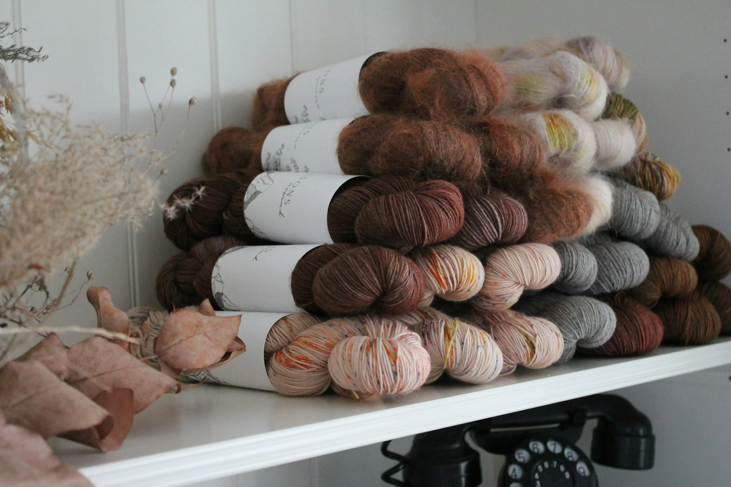 Stacks of multicolored yarn skeins on a white shelf, with dried flowers to the left and a vintage black rotary phone below.