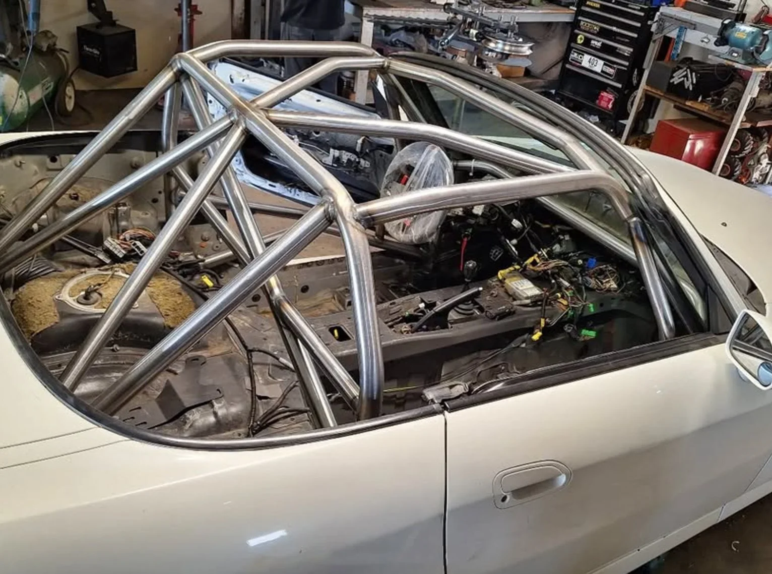 A car in a garage with a metal roll cage frame installed inside, shelf with tools and parts in the background