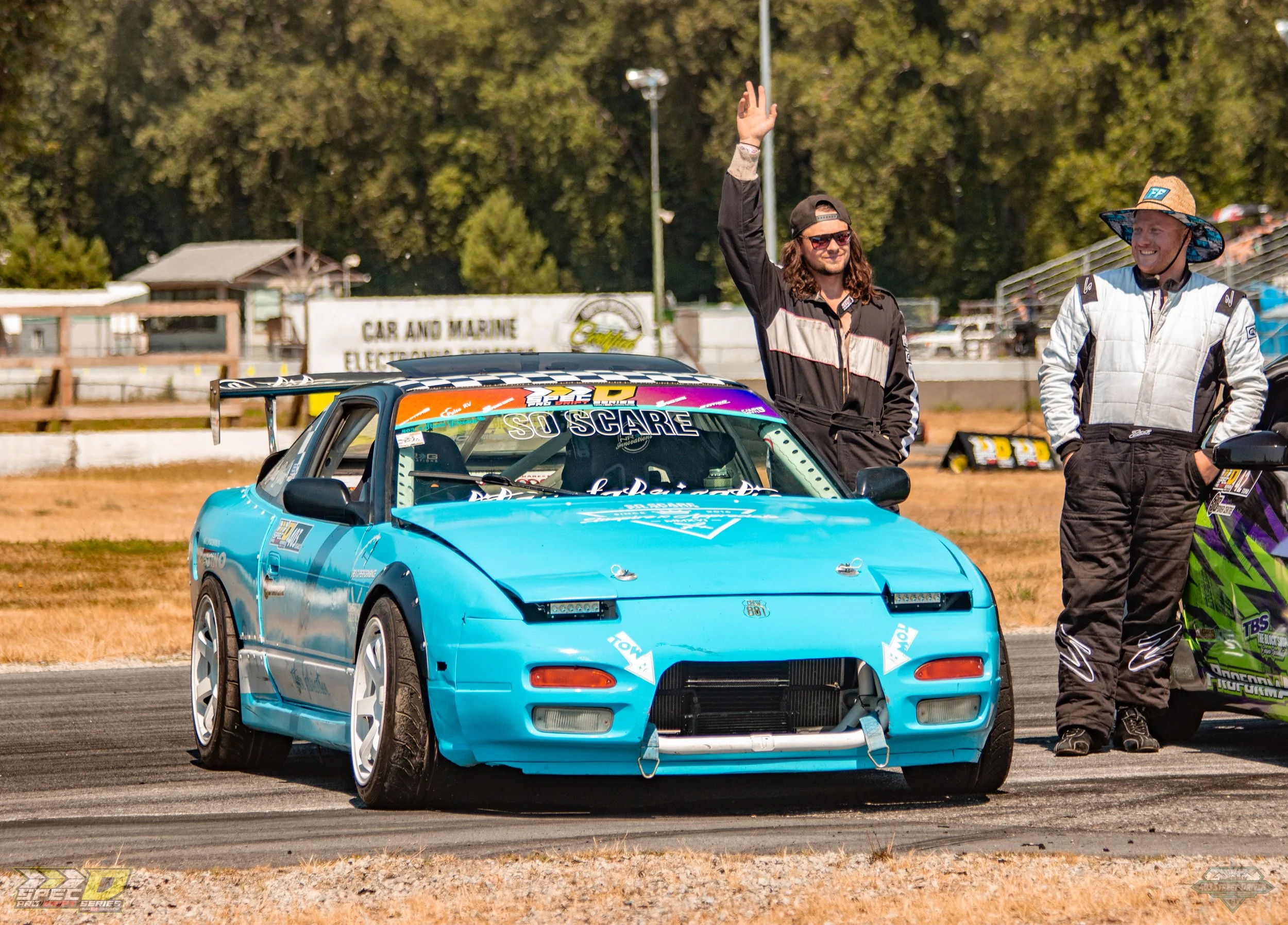 A bright blue race car with a spoiler and white wheels on a track, with two people standing beside it. One person is in a racing suit with their hands in their pockets, the other in a black and white jumpsuit waving with sunglasses and a backwards cap. The background includes trees and a sign that reads 'Car and Marine Electric'.