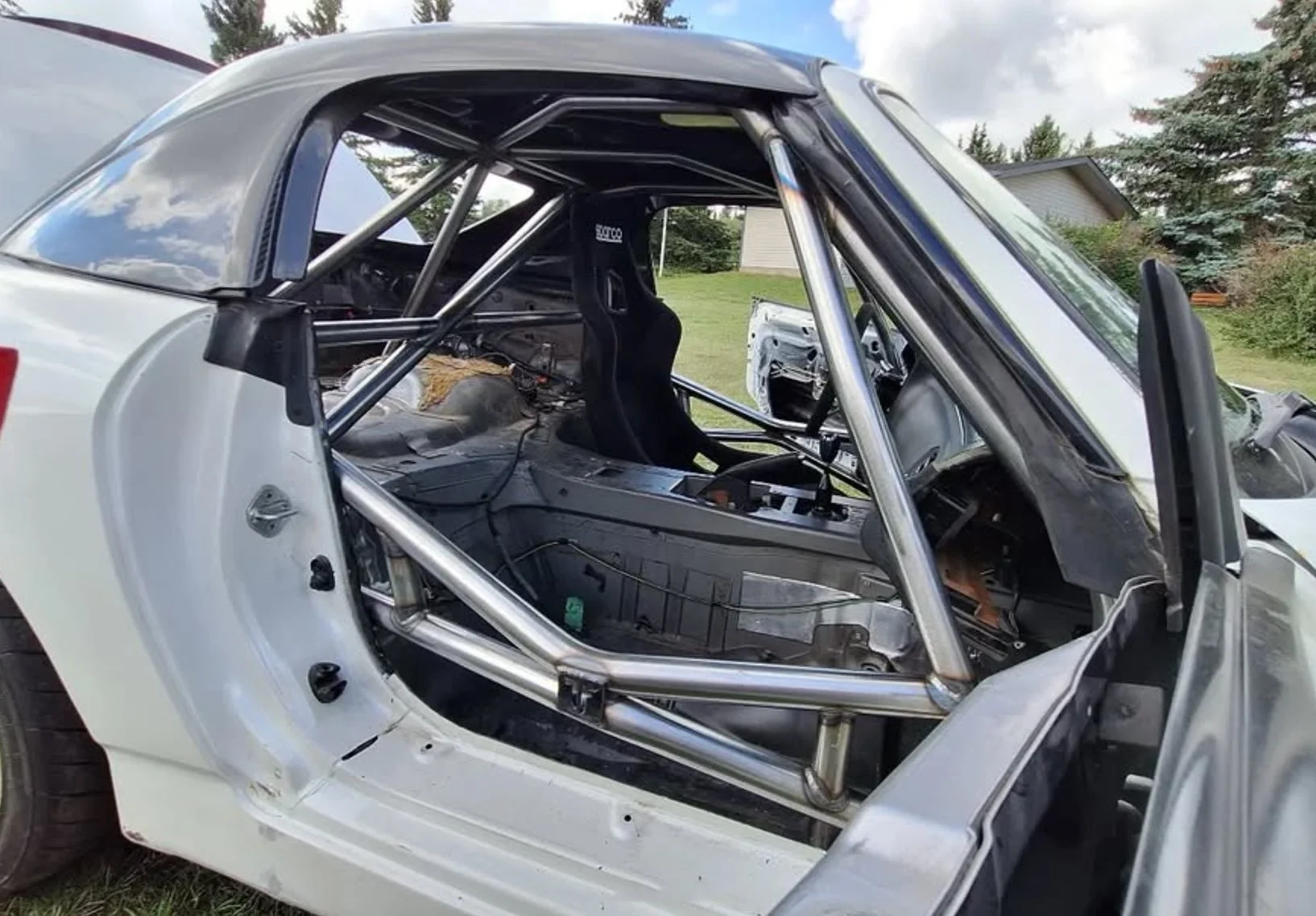 A race car with a silver roll cage inside, no door panels, and a racing seat, parked outside on grass with houses and trees in the background.