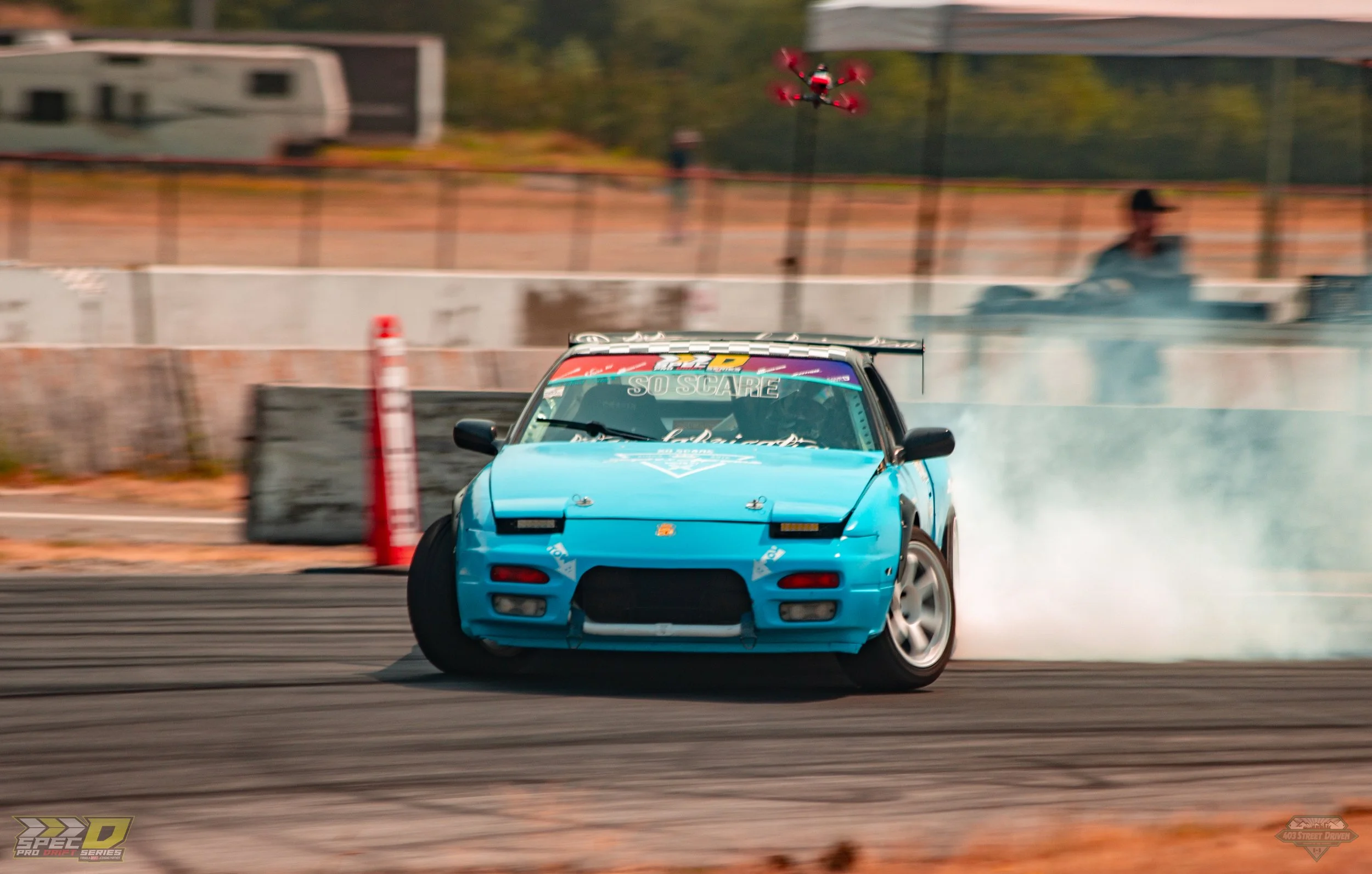 A blue sports car drifting on a race track, producing smoke from the tires, with a drone flying above and a person visible in the background.