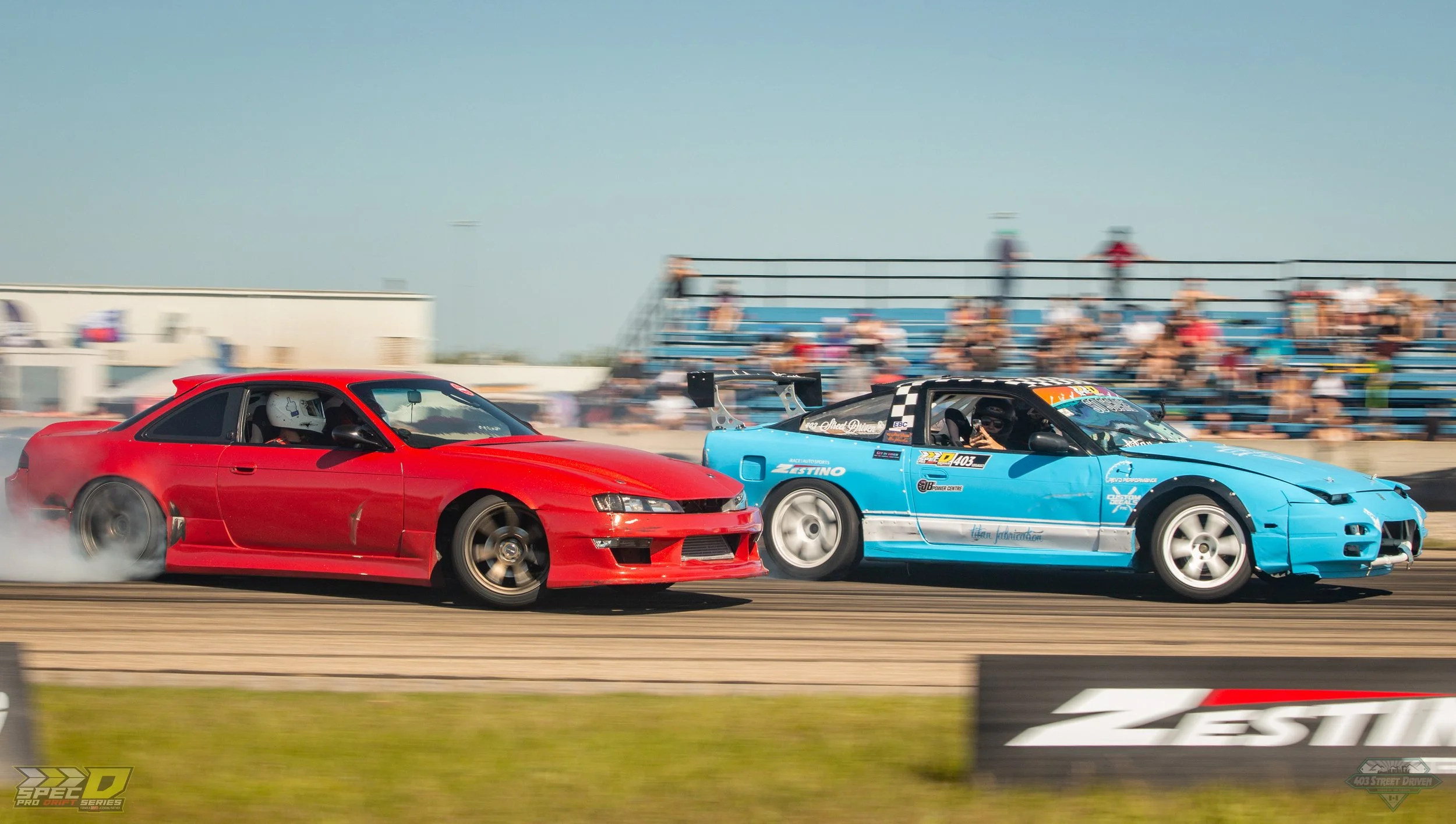 A red sports car and a blue drift car racing on a track, with spectators watching from the bleachers in the background.