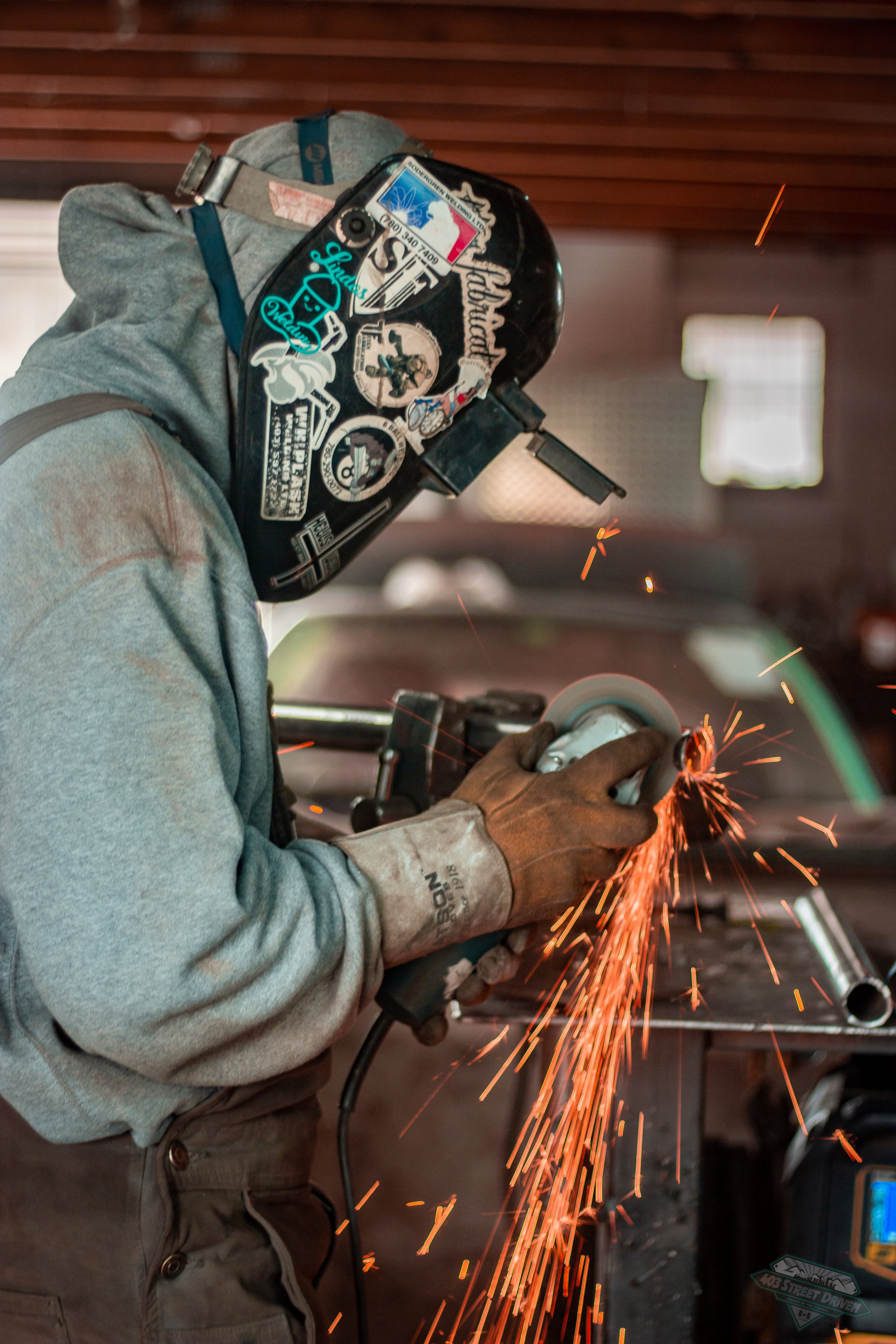 A person wearing a welding helmet and gloves is using a grinder on a metal piece, with sparks flying.